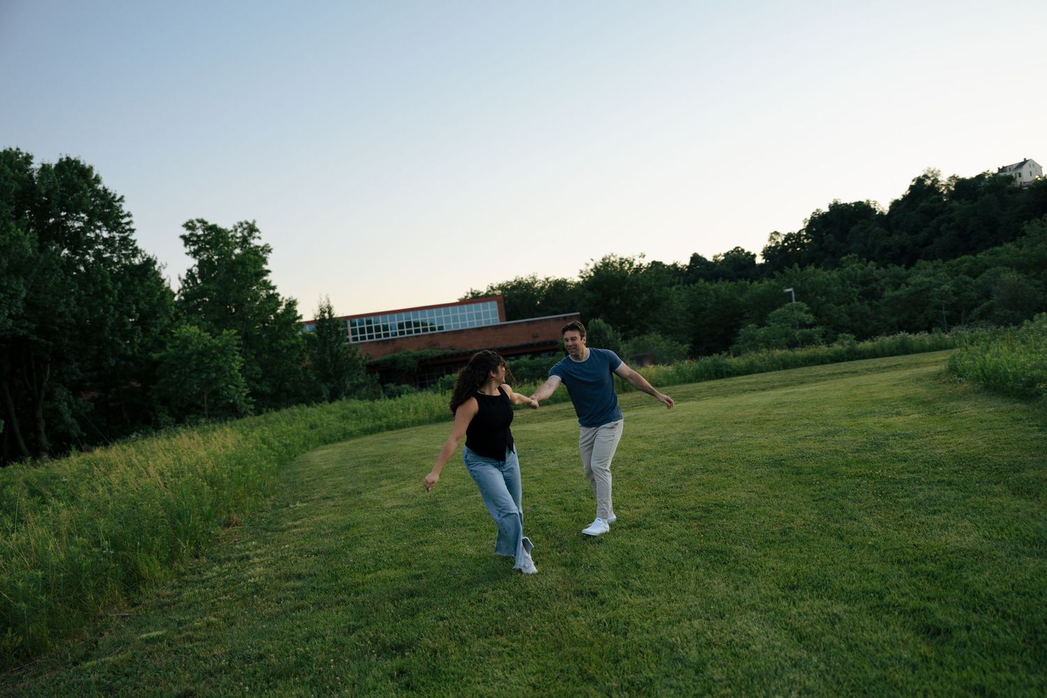 A couple joyfully runs hand-in-hand across a lush green field at dusk, with trees and a building in the background.