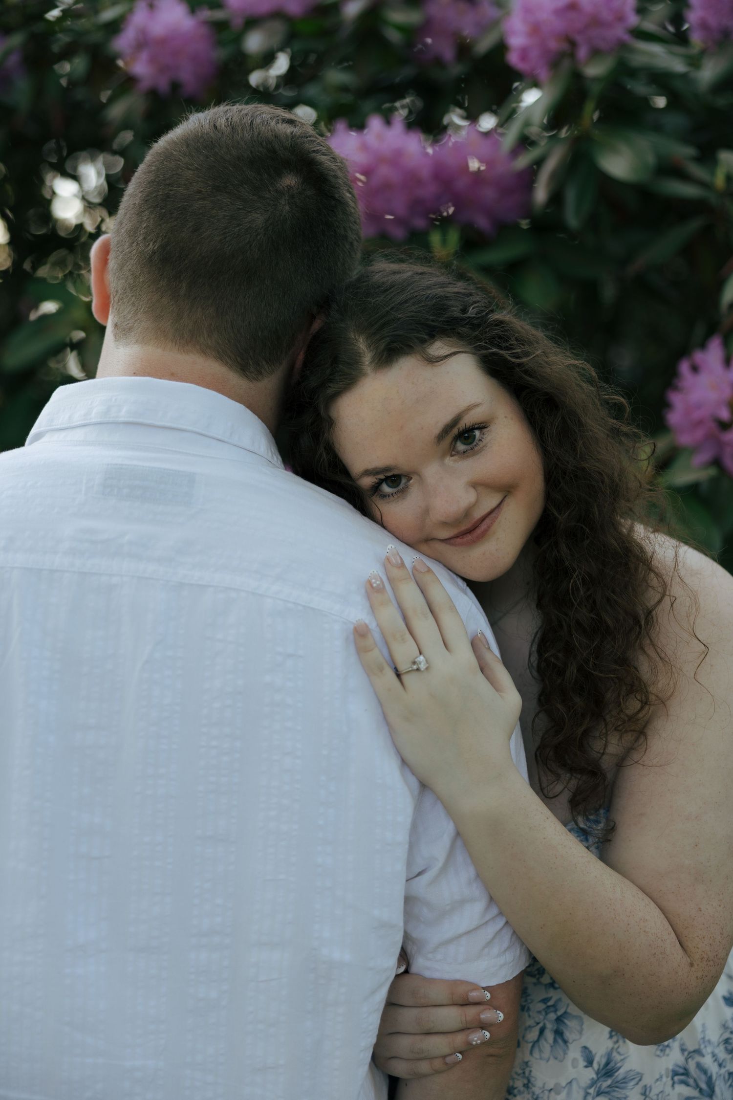 Woman with curly hair smiling over partners shoulder while embracing near vibrant pink flowering shrubs.