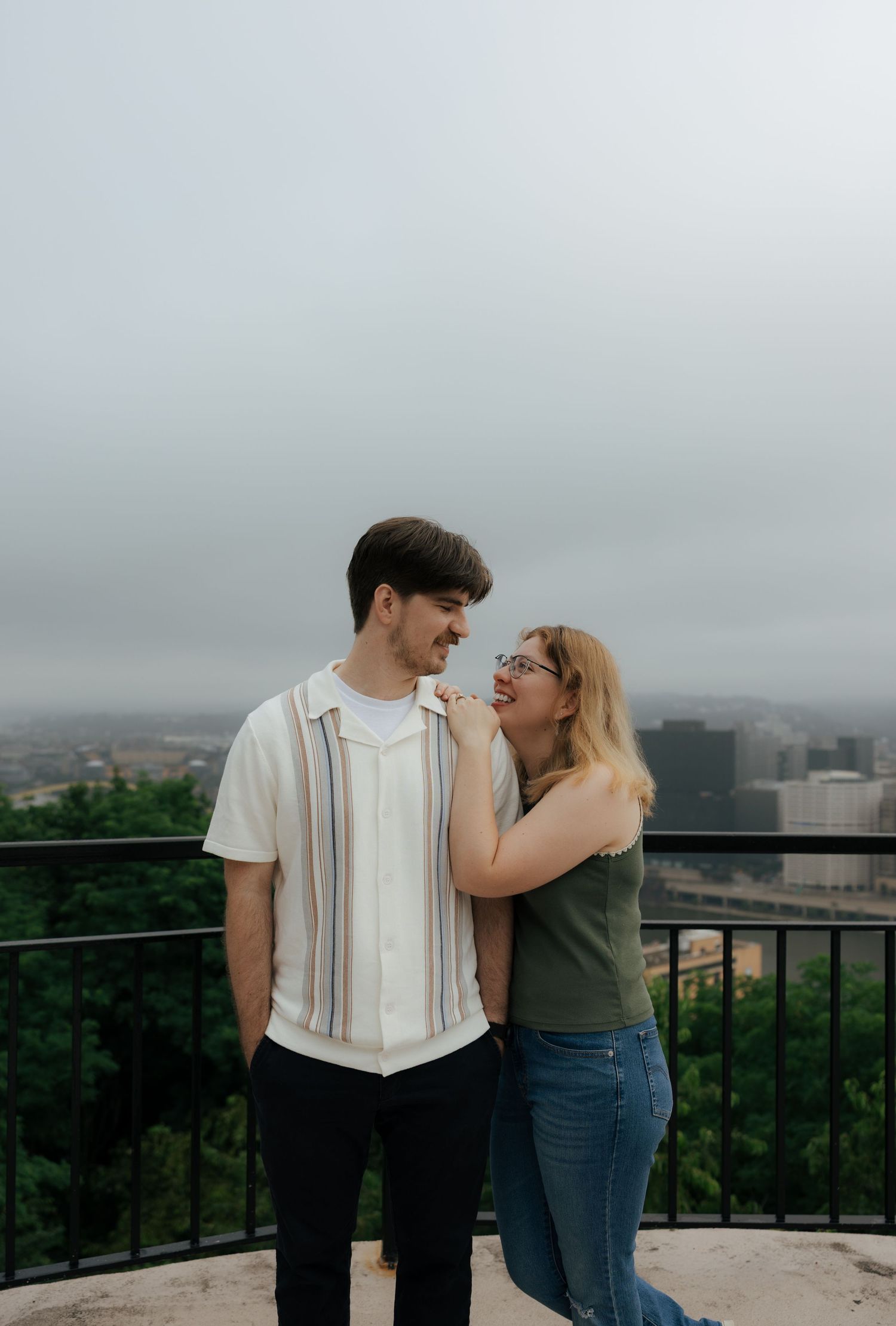 Couple smiling at each other on an overlook with a foggy city skyline behind them.
