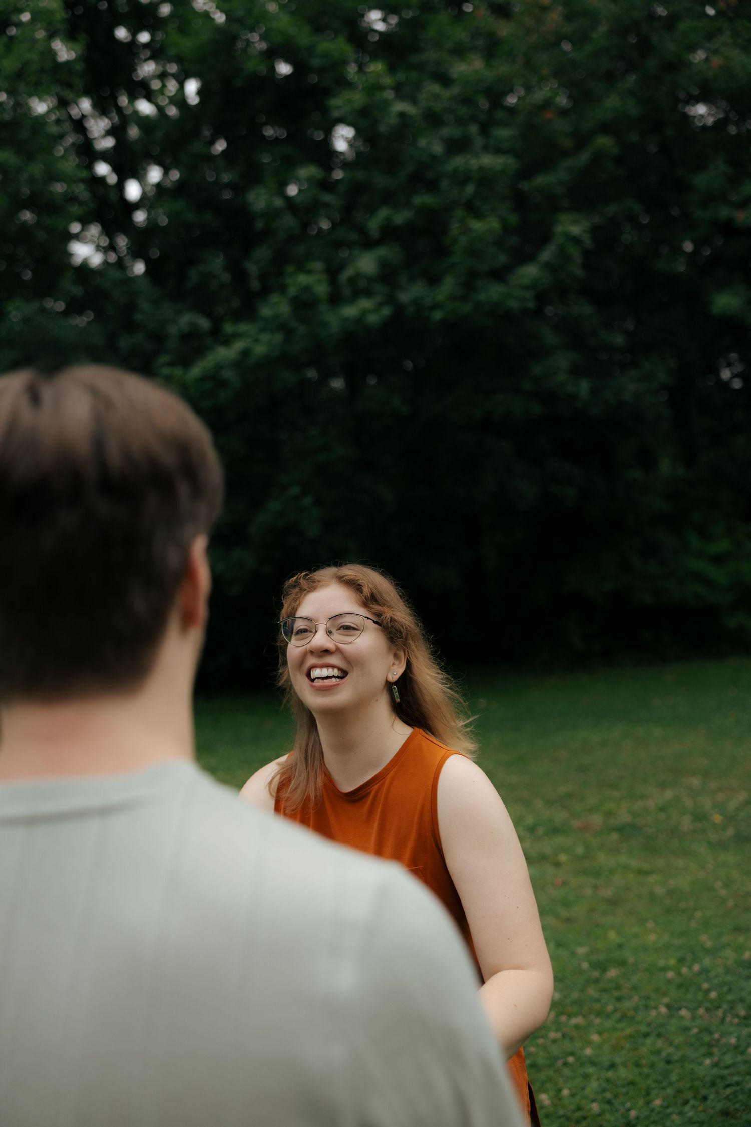A woman with glasses laughs joyfully while facing a man in a green outdoor park setting.