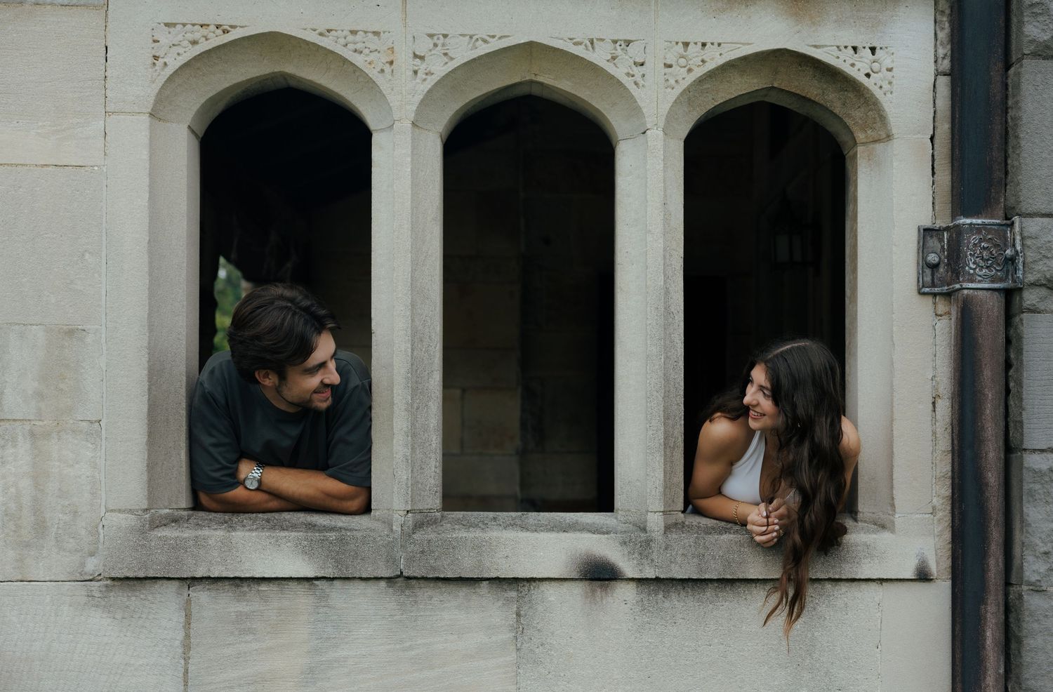 A man and woman lean out of arched stone windows, smiling at each other on a historic building facade.