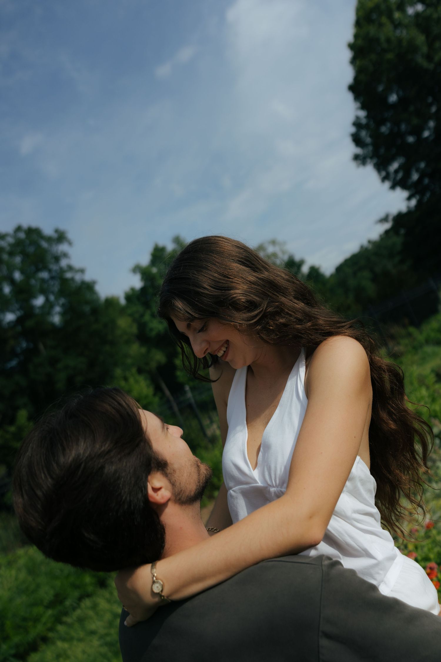A couple gazes at each other lovingly outdoors, the woman smiling brightly while seated on the man's lap.