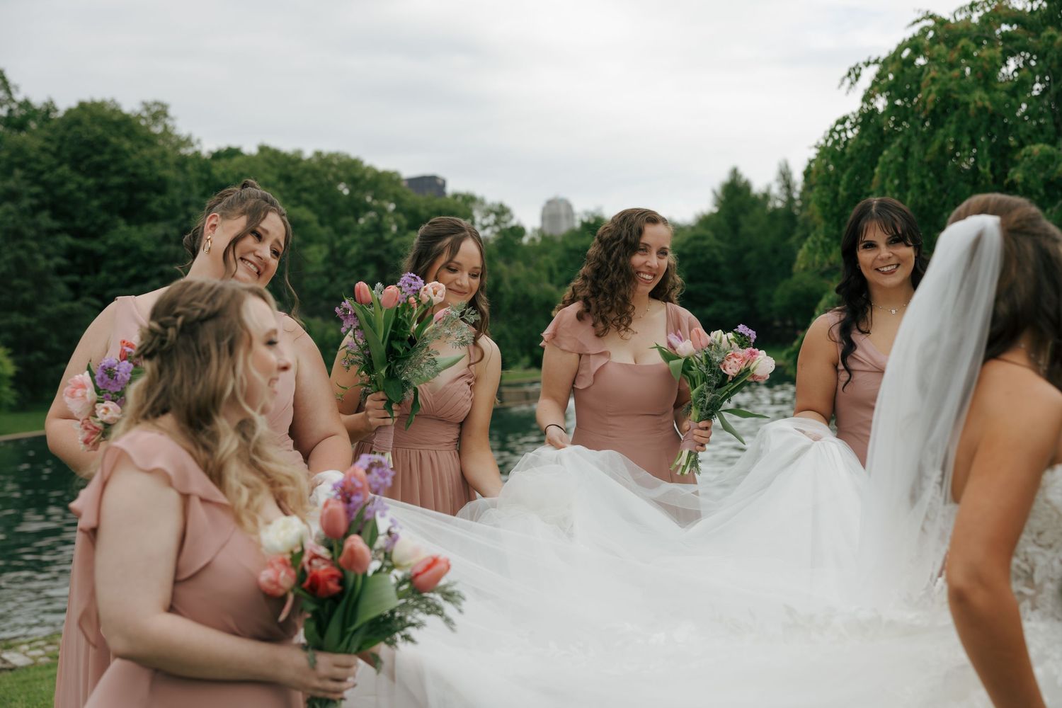 Bride in white gown with bridesmaids in pink dresses holding colorful tulip bouquets near a pond in Central Park.