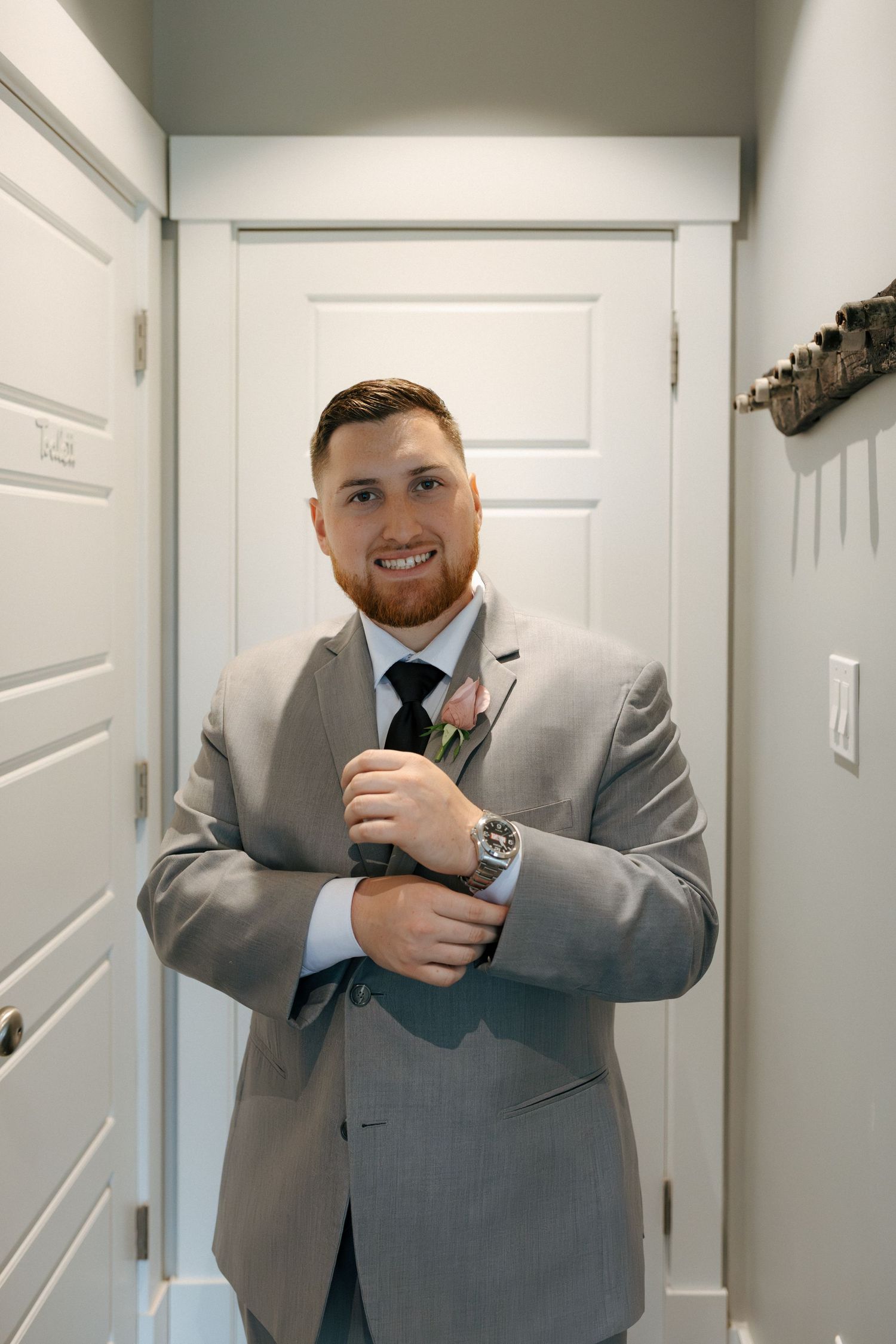 Smiling groom in grey suit adjusting cufflinks with pink boutonniere and black tie before wedding ceremony.