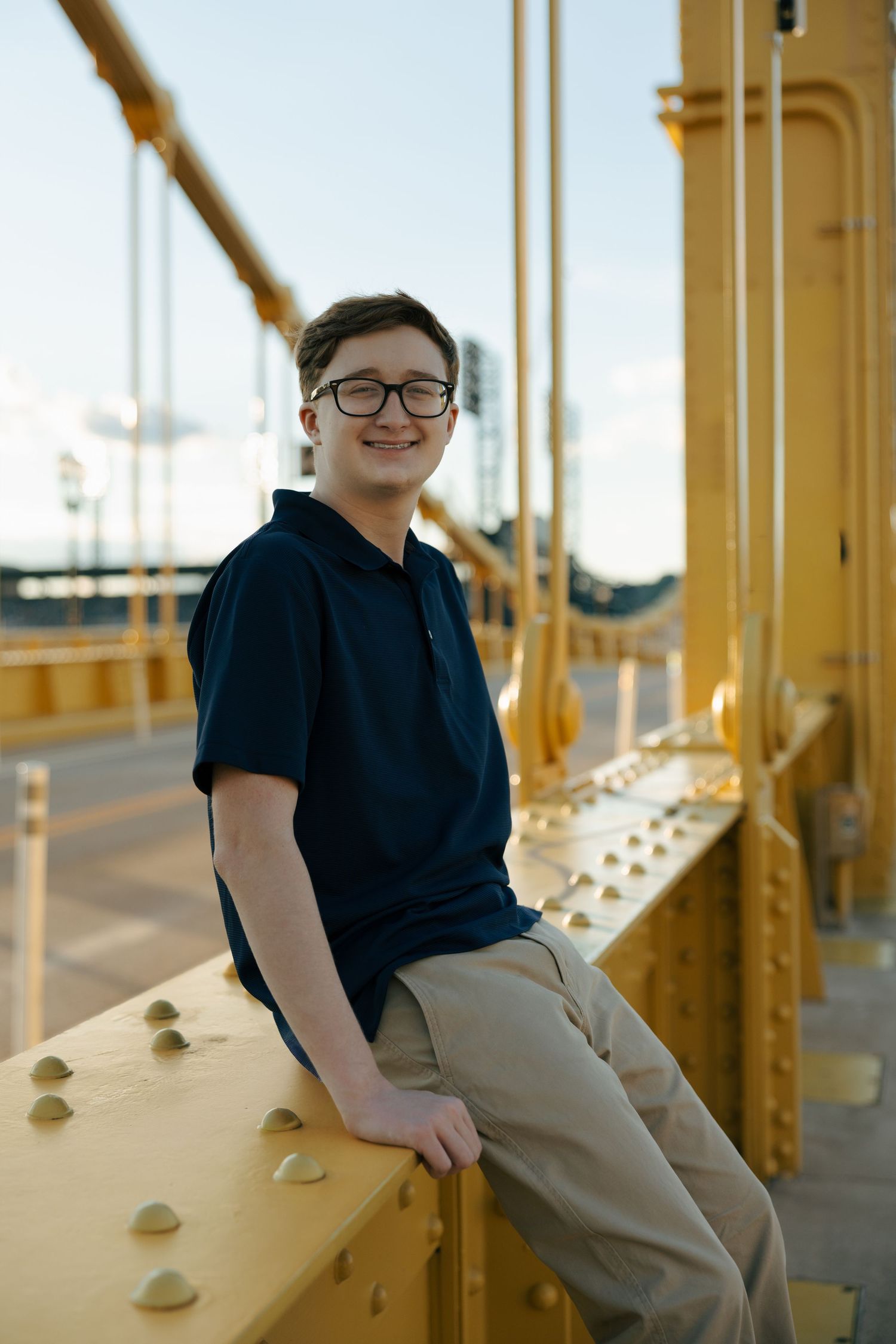 Young man in glasses and navy polo sitting on yellow steel bridge structure against blue sky.