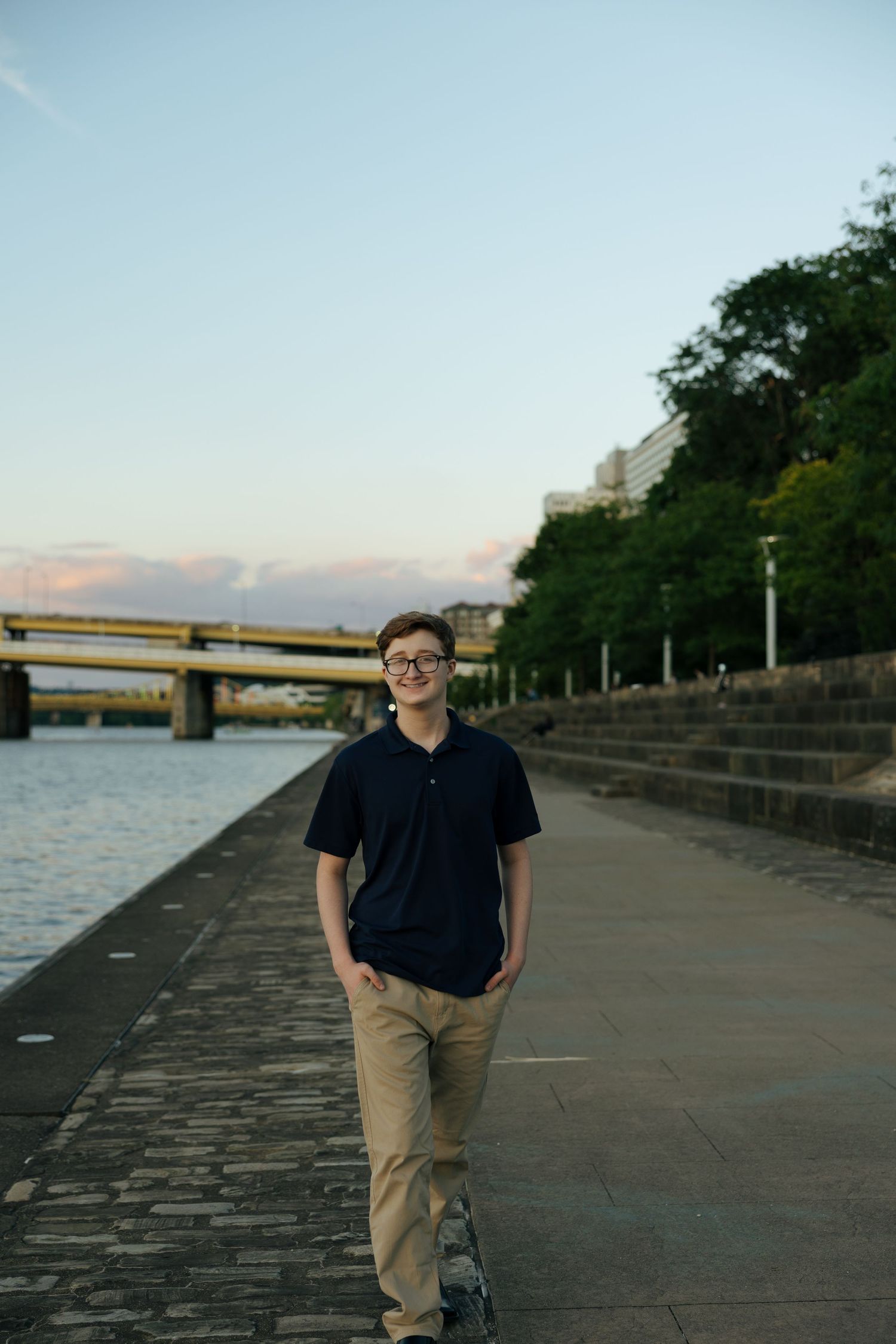 Teen boy in navy polo and khakis walking along urban riverfront path at dusk with bridge view.