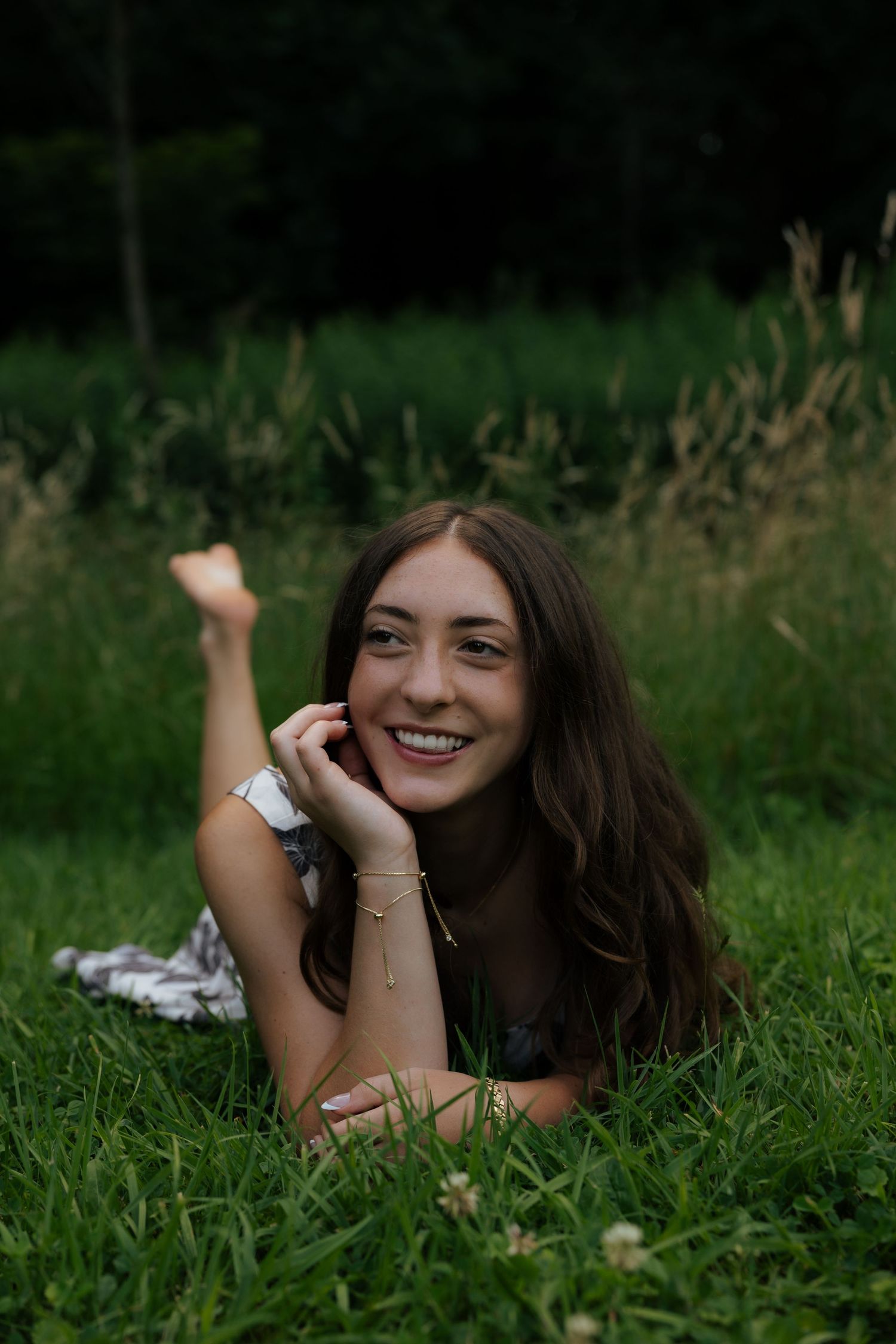 Young woman with long brown hair lying on green grass, smiling brightly, wearing bracelets, with wildflowers nearby.