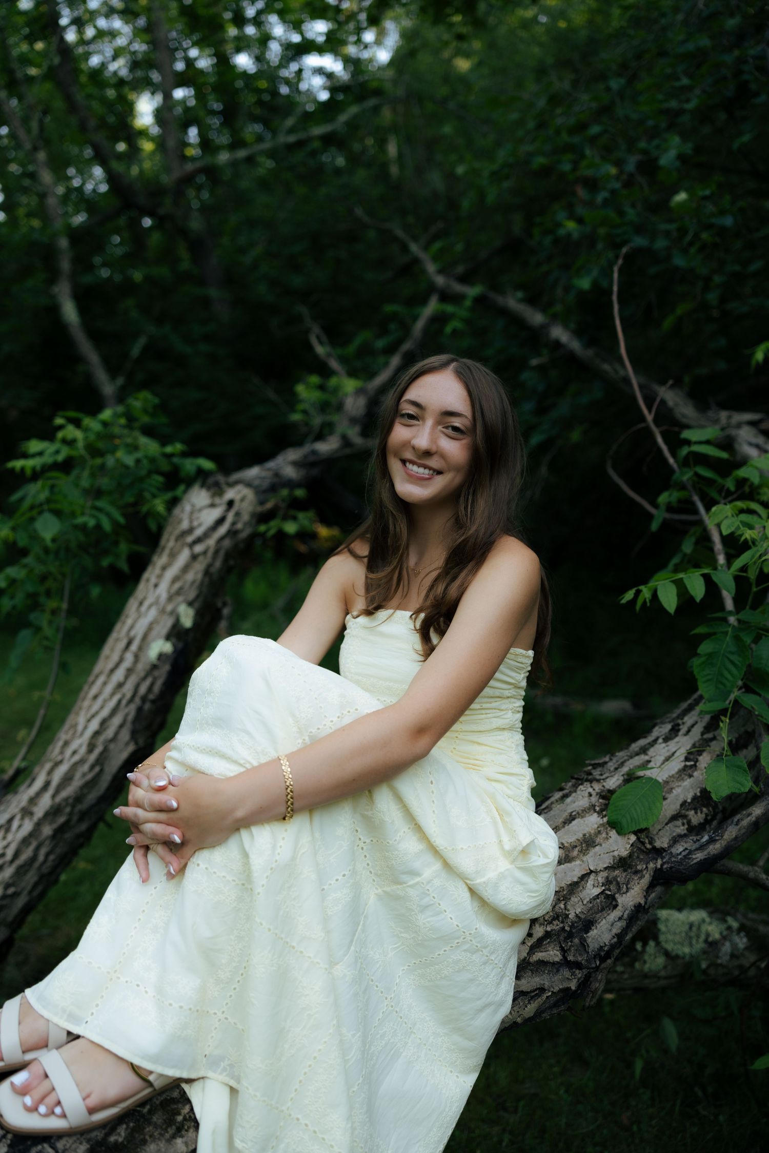Happy young woman in a white strapless gown sitting on a fallen log surrounded by lush green trees.