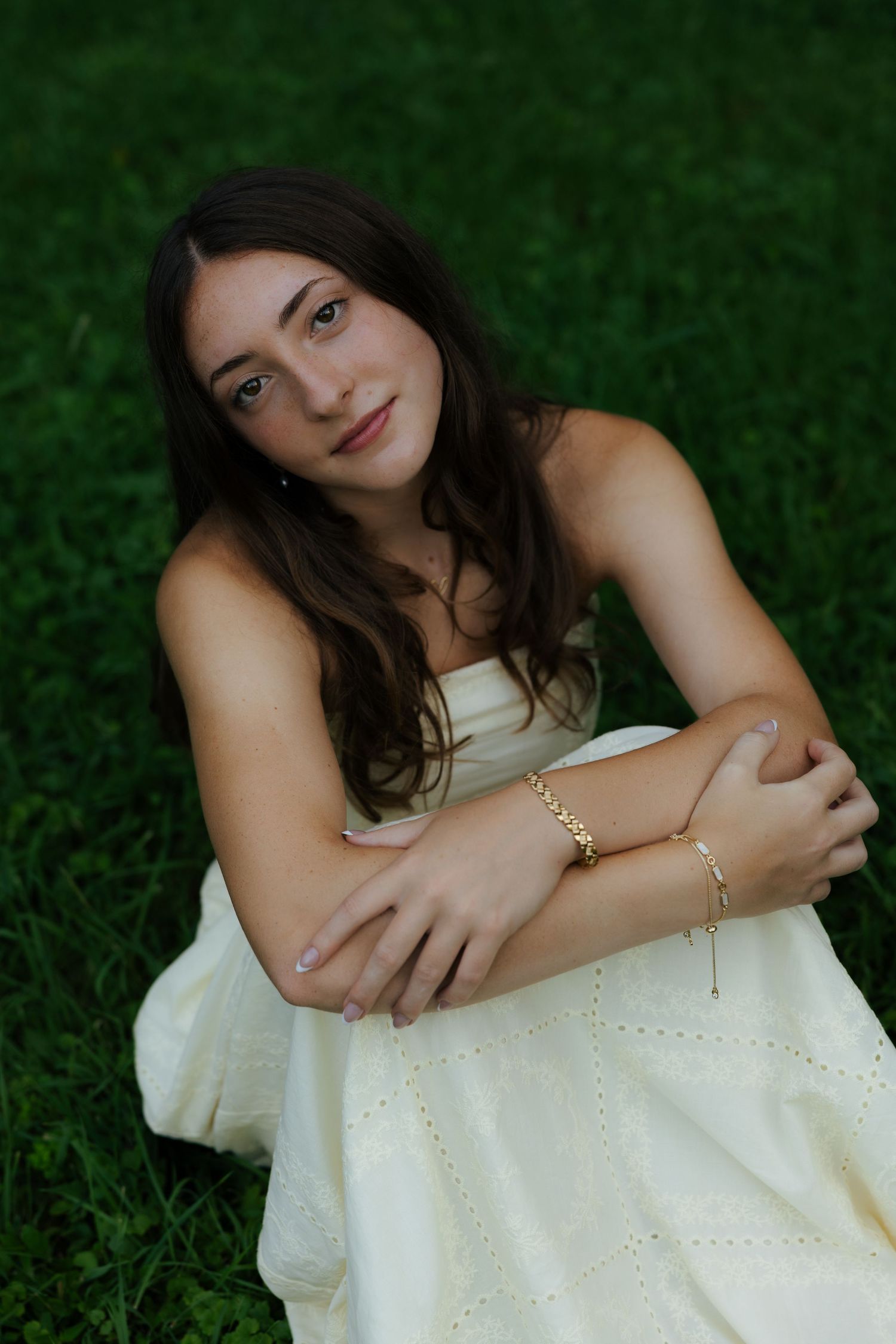 Young brunette woman in cream strapless dress sitting on grass with gold bracelets smiling.