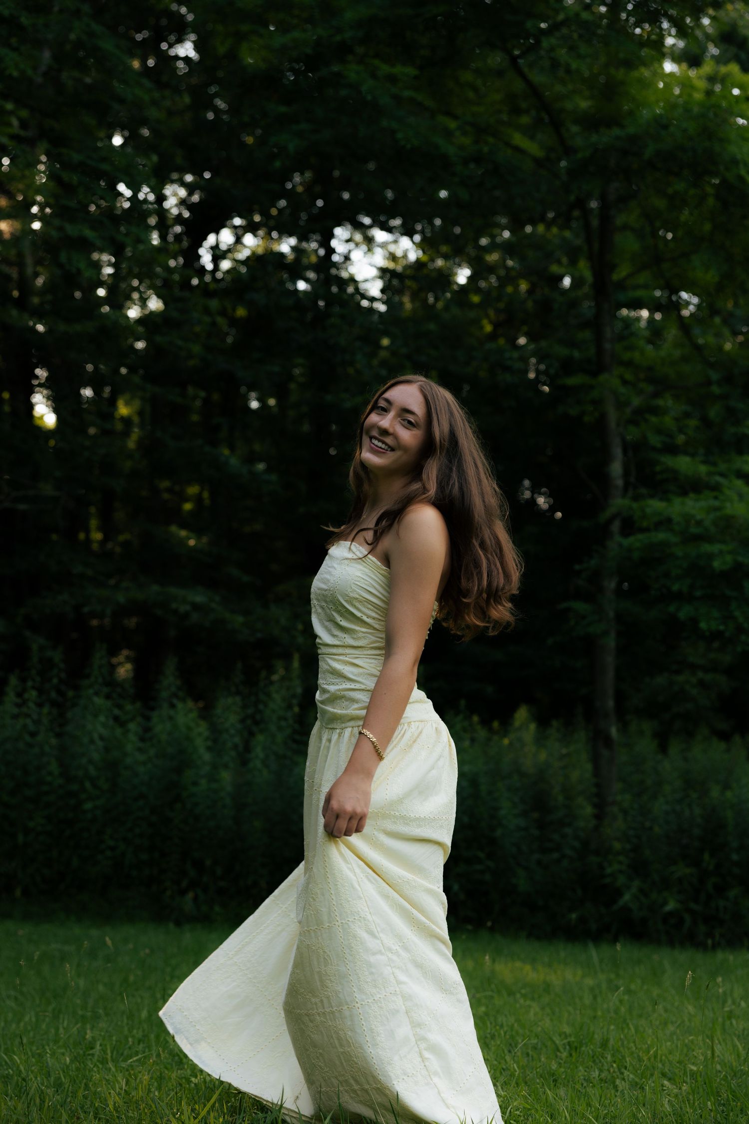 Young woman in flowing yellow dress twirling and smiling in lush green garden at dusk.