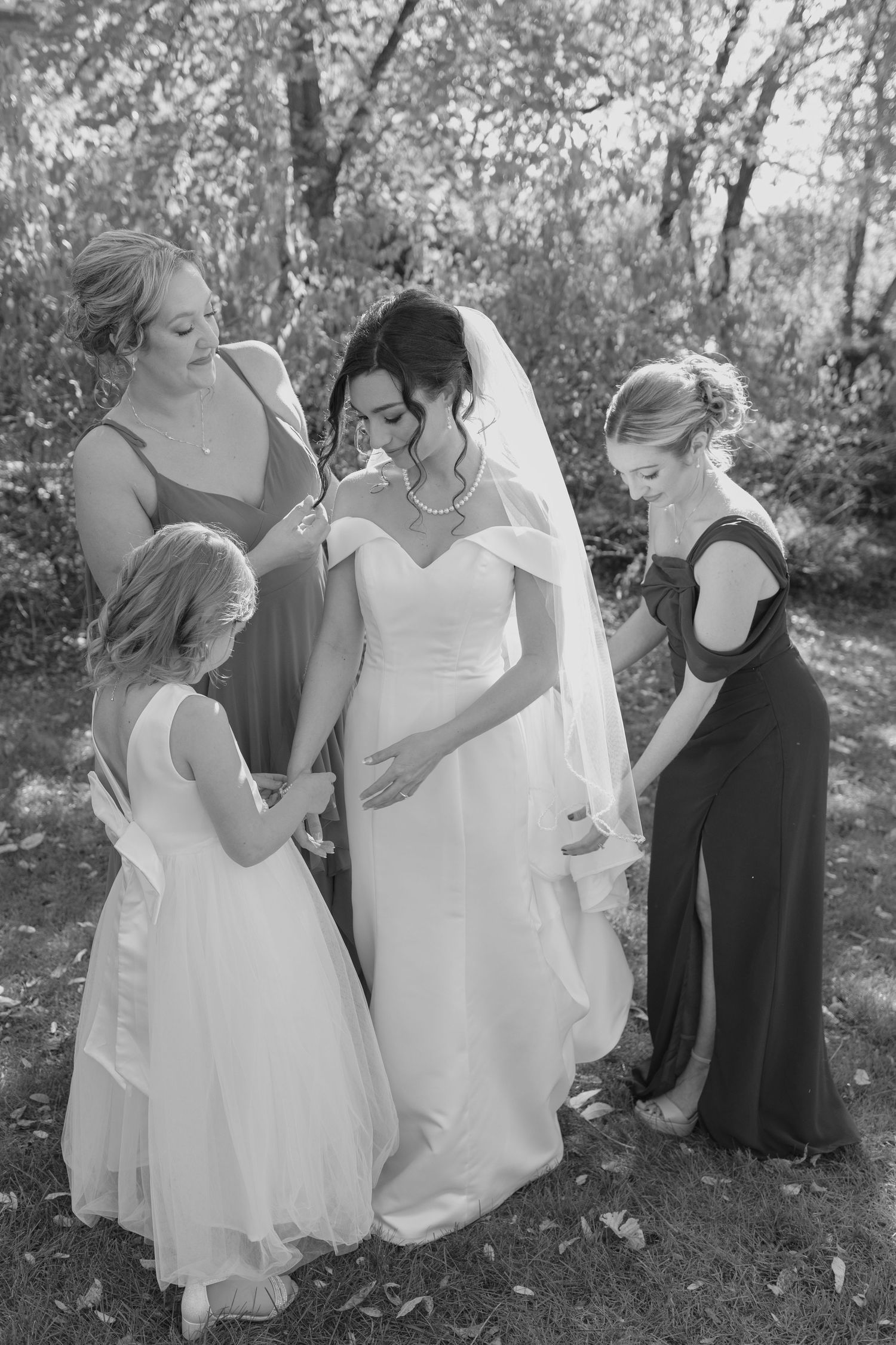 Bride in off-shoulder gown shares a sweet moment with bridesmaids and flower girl in a black and white photo.