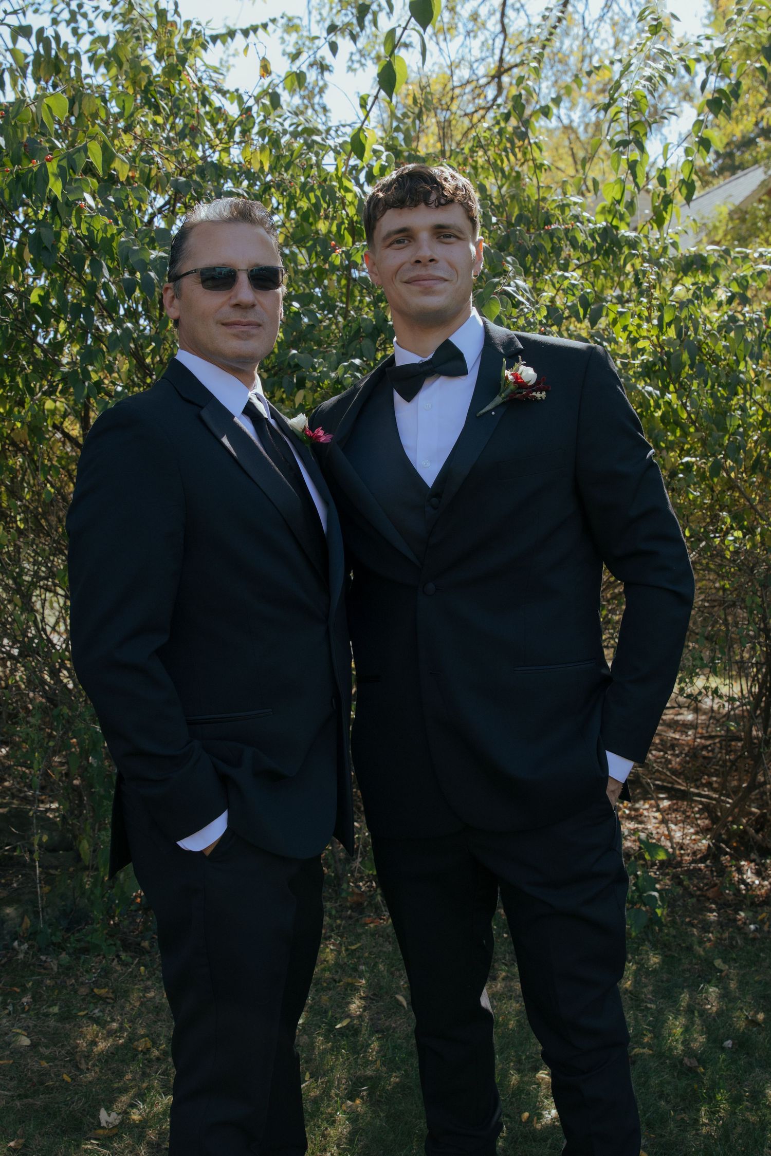 Two men in black suits posing together outdoors, one wearing sunglasses.