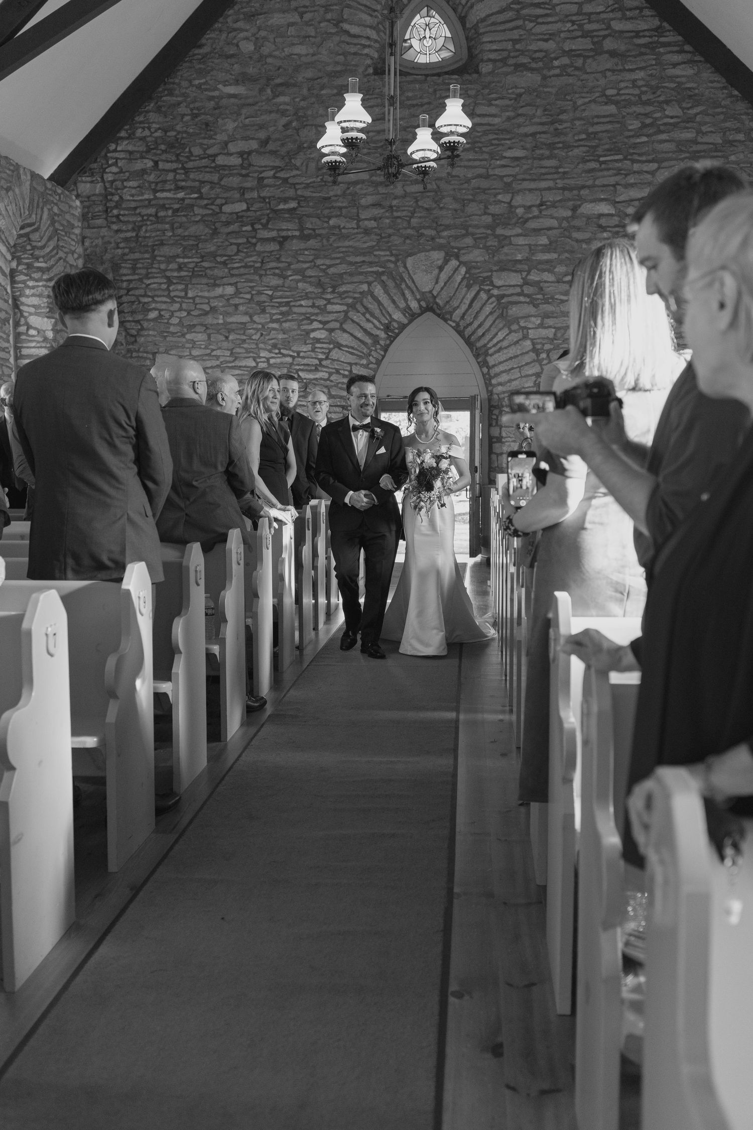Black and white photo of a bride walking down the aisle in a stone chapel with guests standing in pews.
