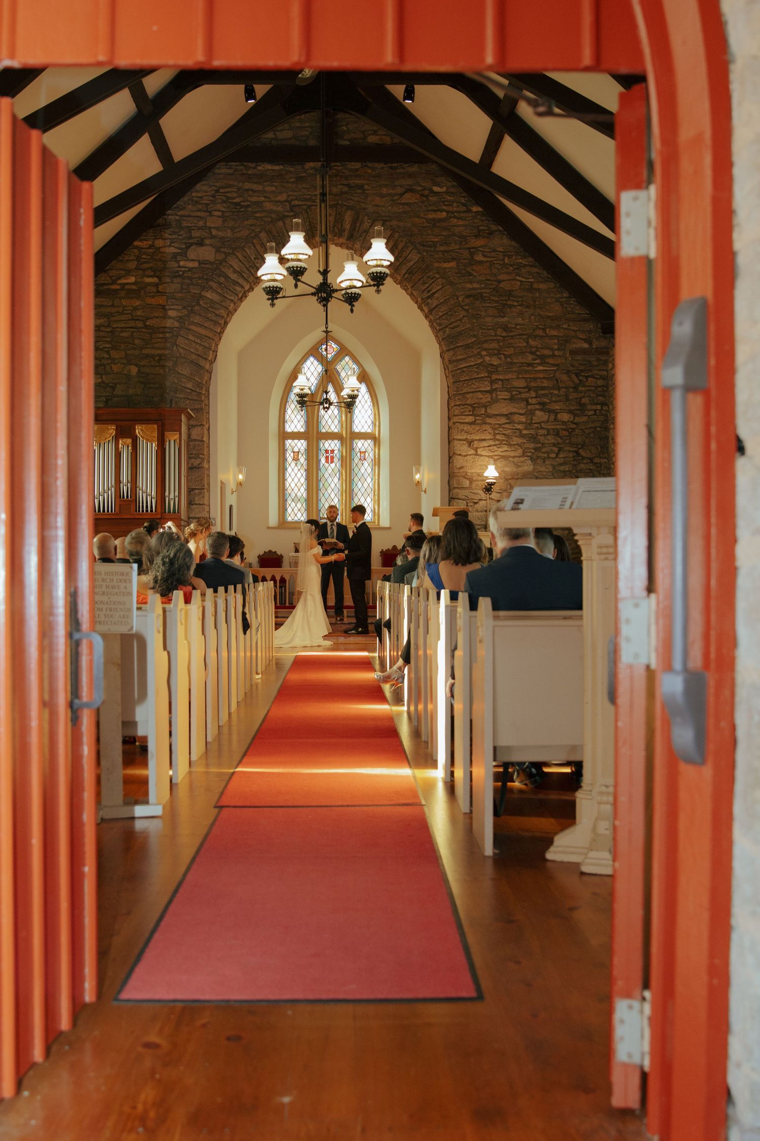 A wedding ceremony viewed through open red doors of a stone church with a red carpet aisle.