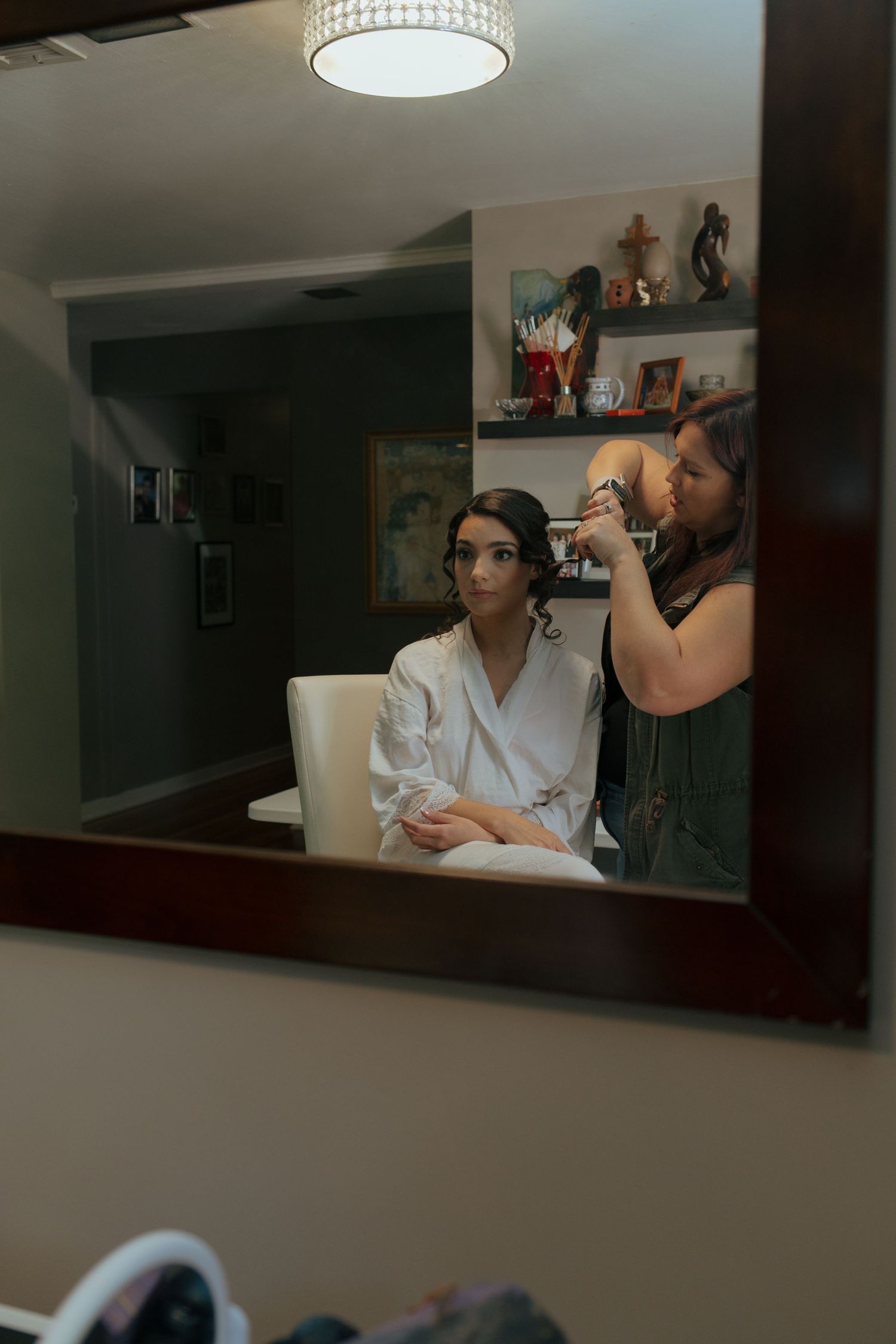 Bride getting hair done by stylist, reflected in ornate mirror during wedding preparation in a cozy room.