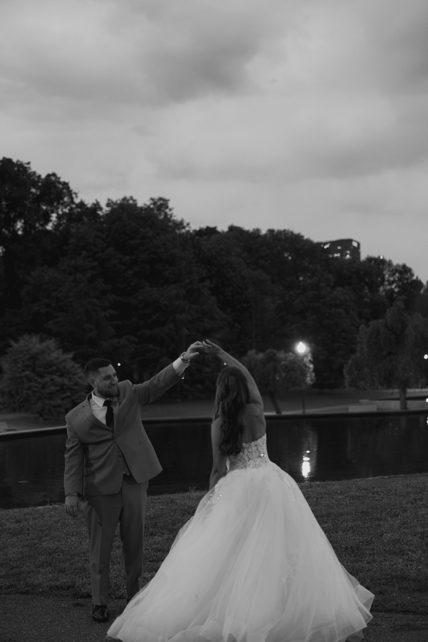 Black and white photo of groom spinning bride in tulle gown by a lake at twilight.