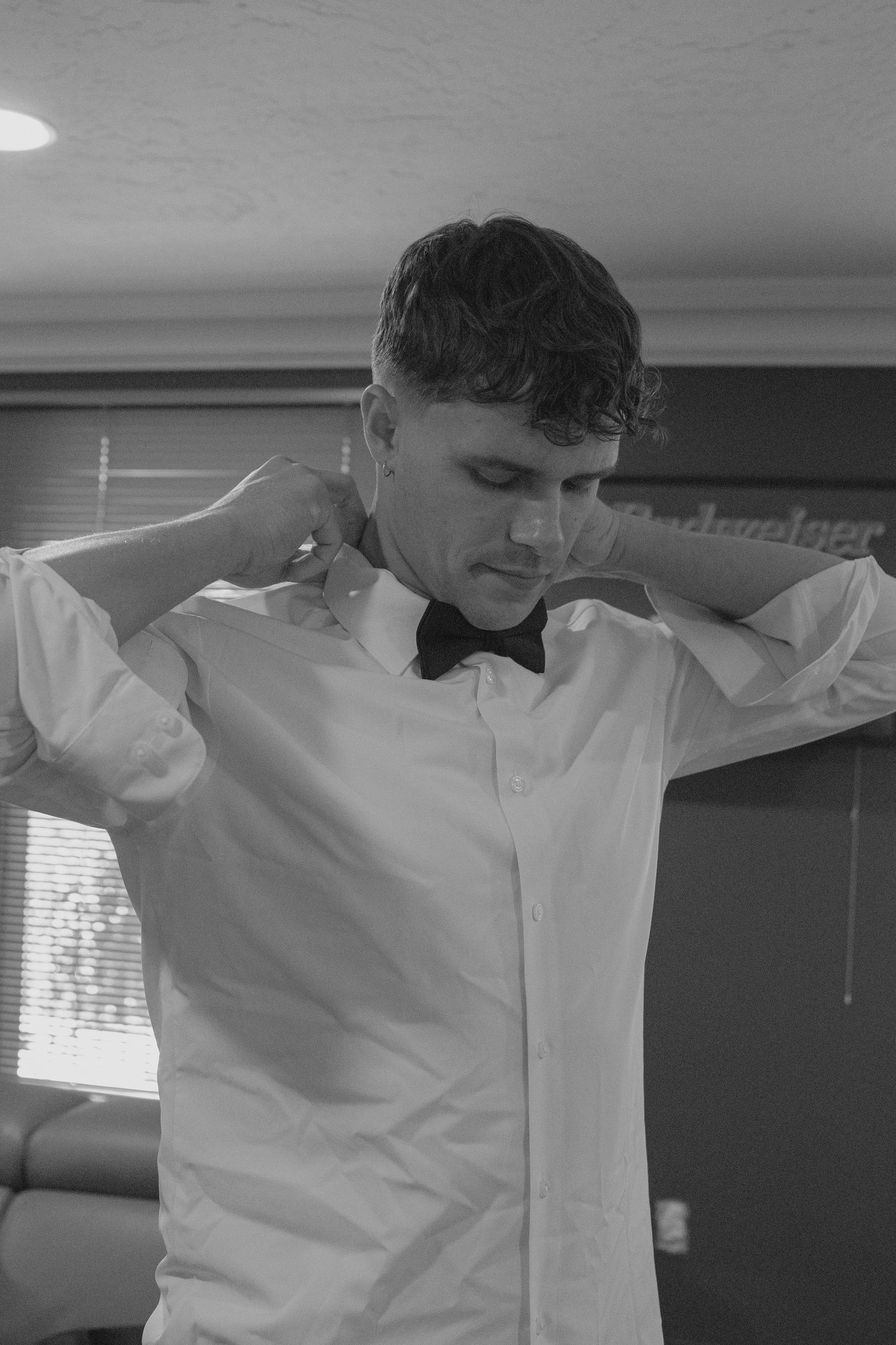 Groom in white shirt and black bow tie adjusting his collar in black and white photo.