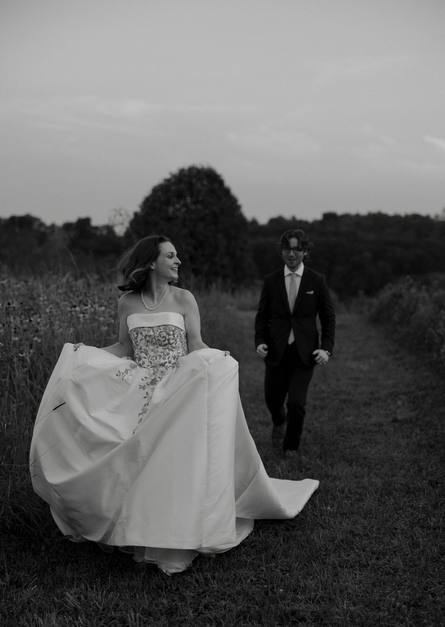 Black and white photo of bride and groom walking along a field path at dusk, bride\u2019s gown flowing in the breeze.