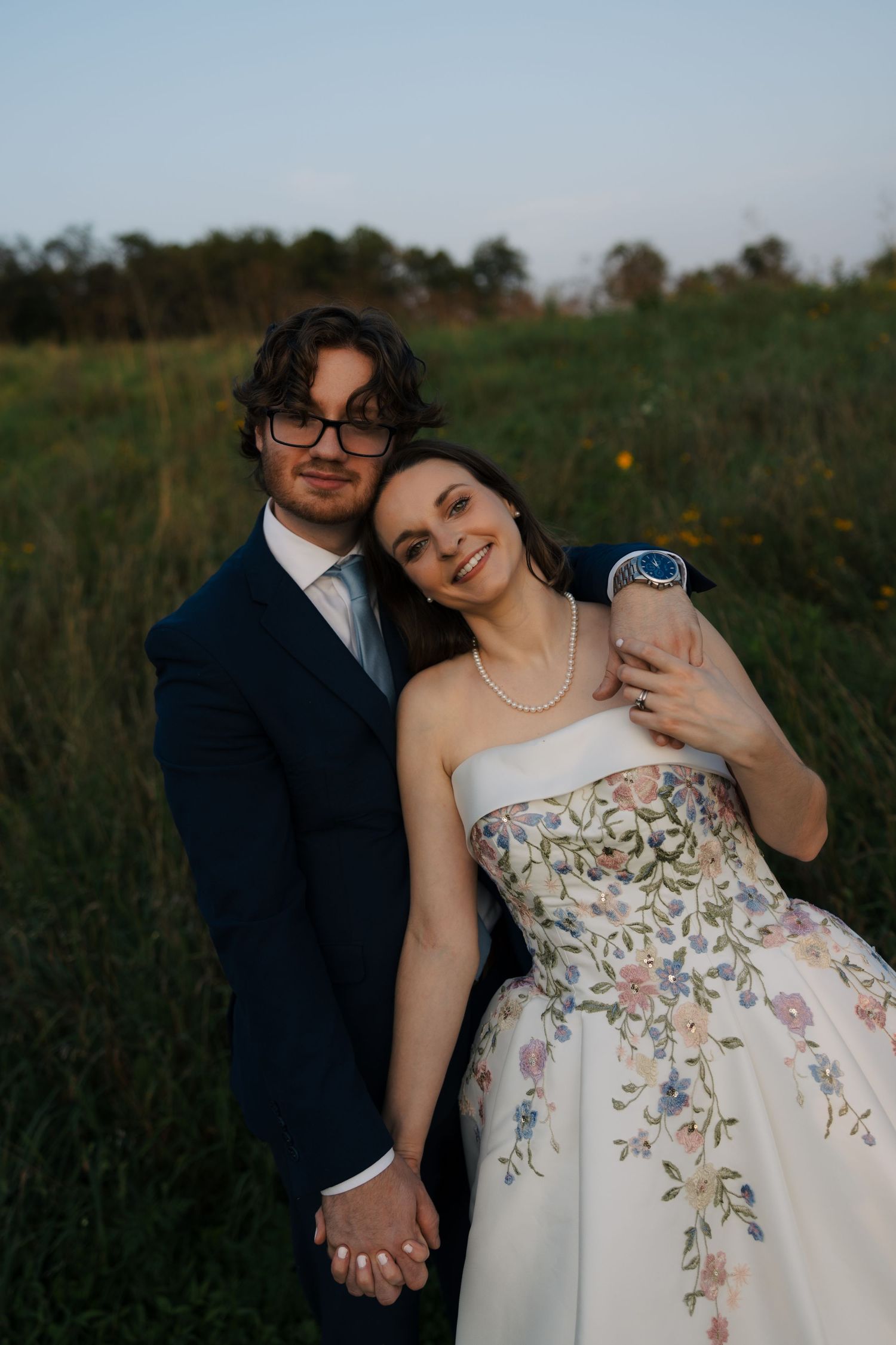 Bride in embroidered floral gown leans against groom in navy suit holding hands in green field.