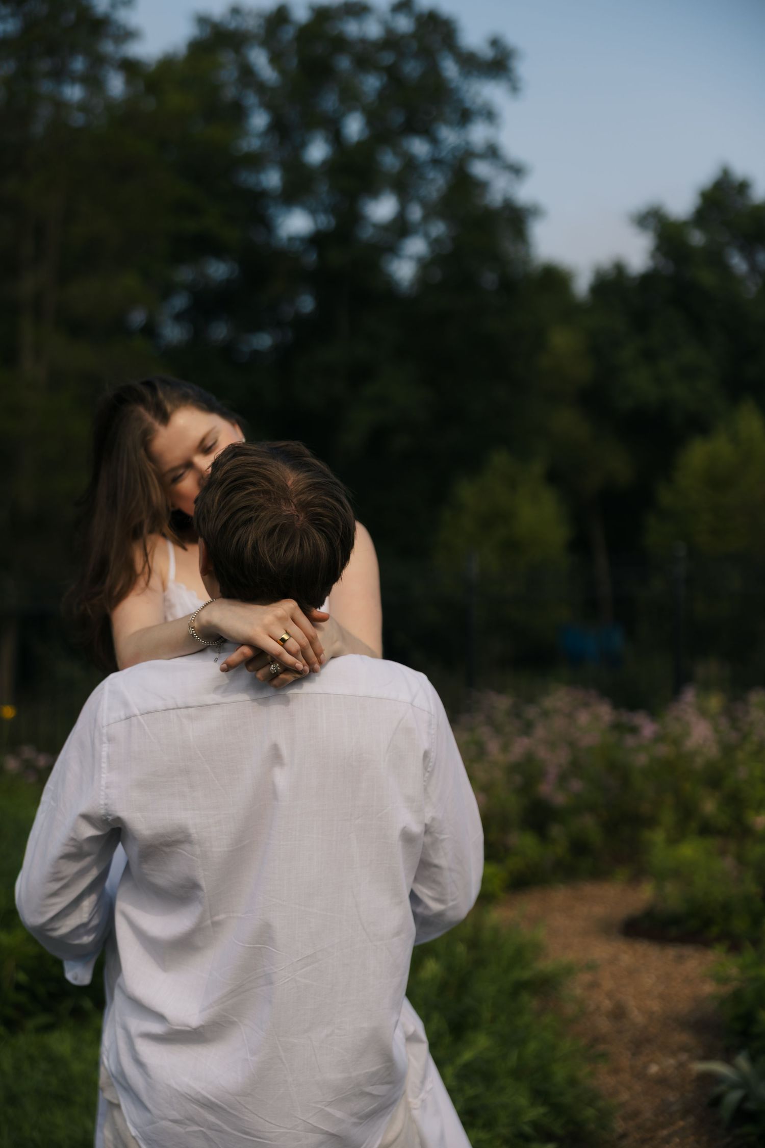 Woman embracing man from behind in a garden path, both wearing white, surrounded by lush greenery.