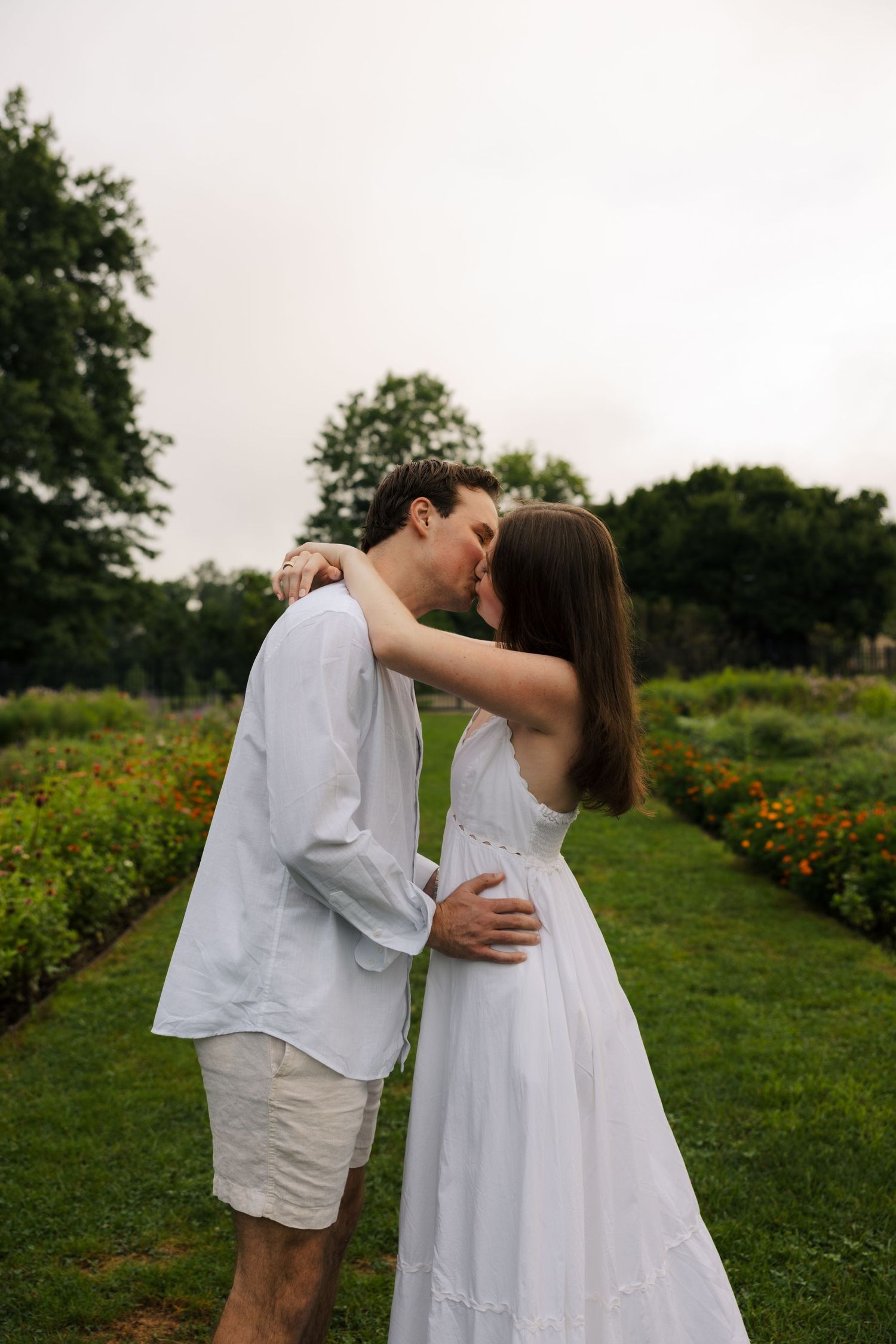 A couple shares a romantic kiss in a lush garden, dressed in white summer outfits, surrounded by colorful flowers.