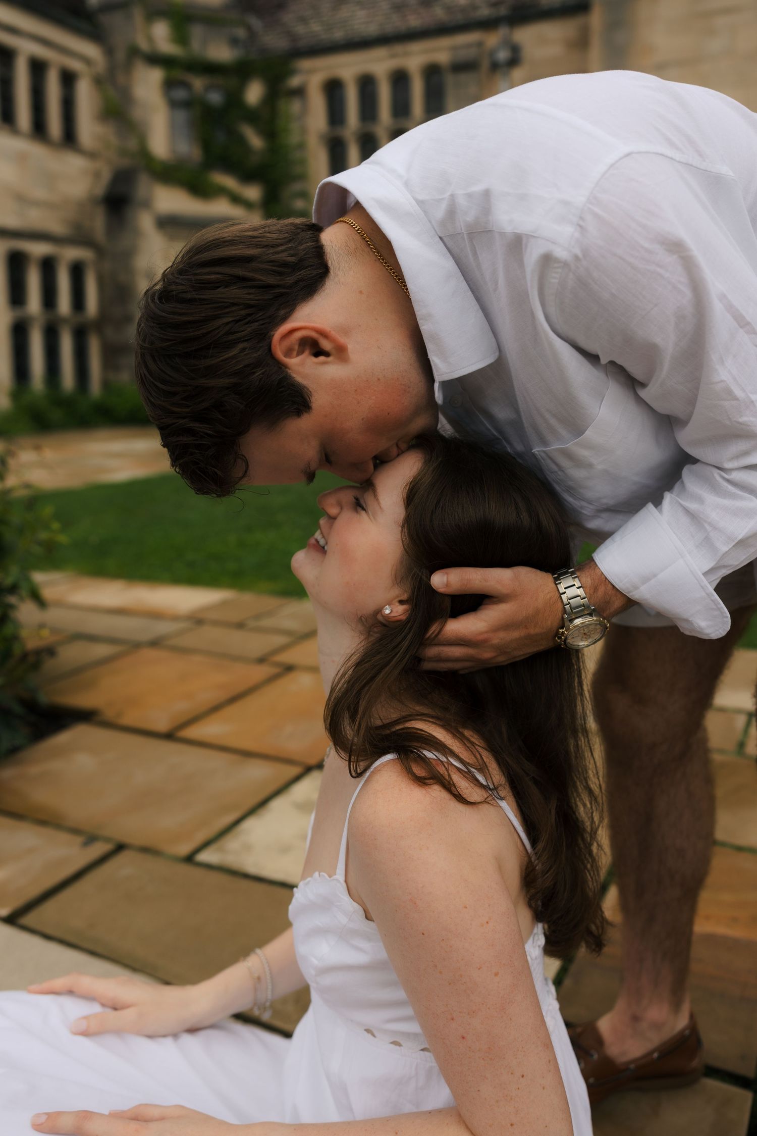 Tender moment as man gently kisses womans forehead while she sits on stone patio outdoors.