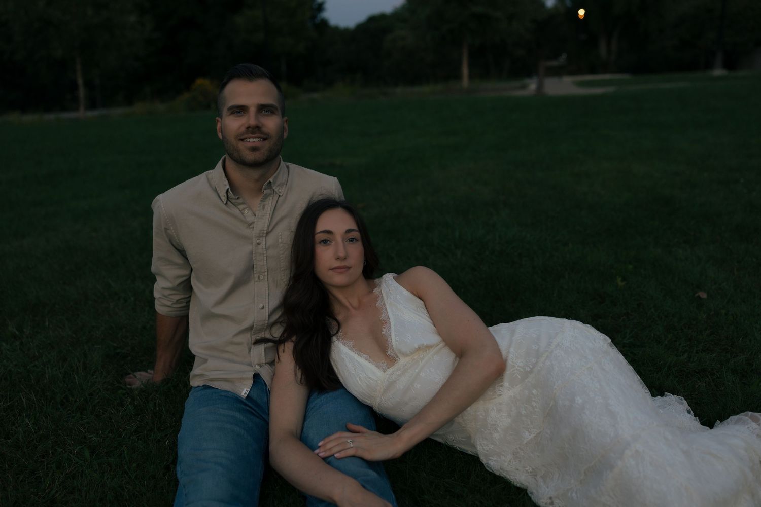 Engaged couple poses on green lawn at dusk, woman in white dress leaning against partner.