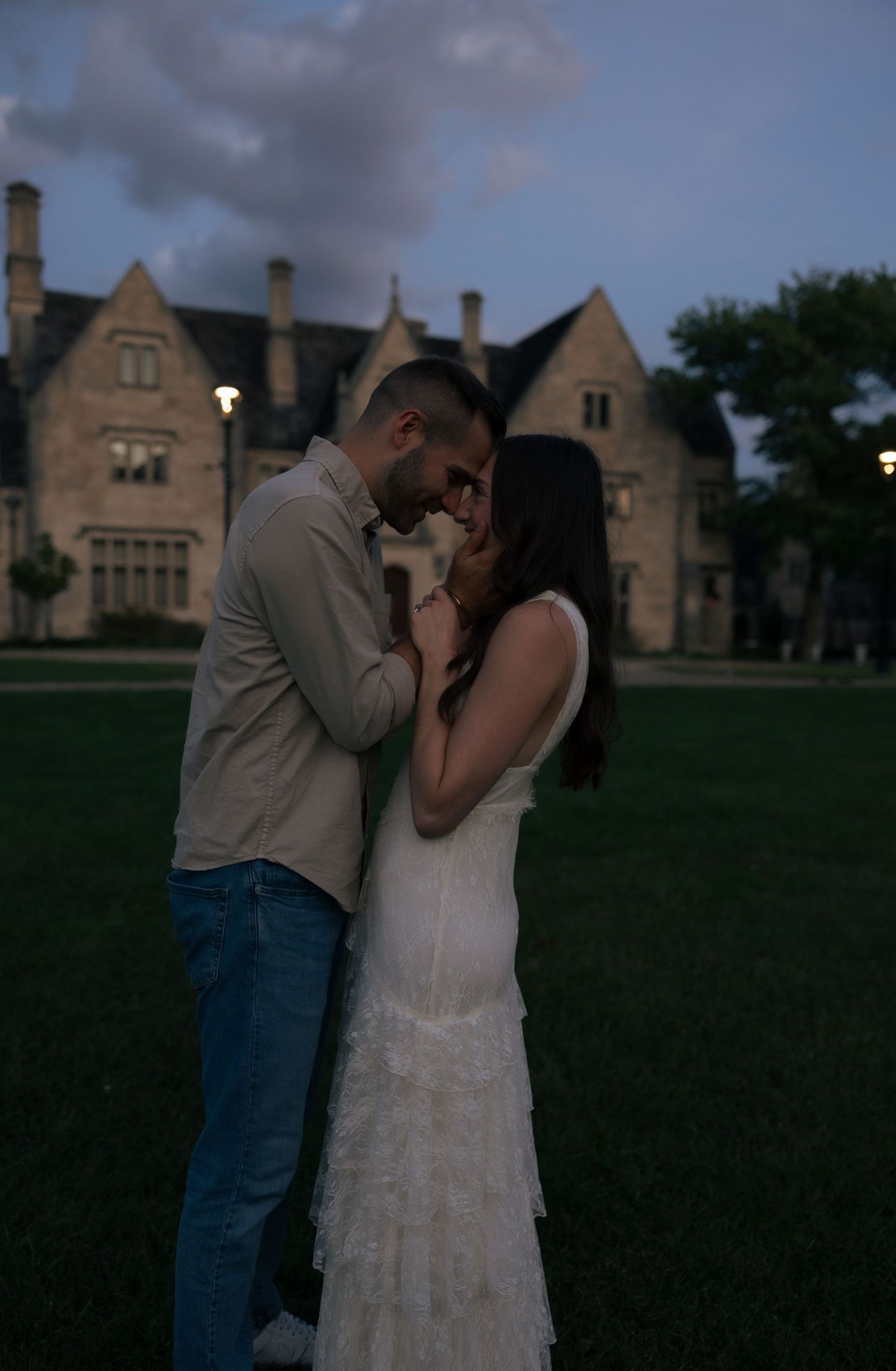 Couple sharing a romantic moment at dusk in front of a grand stone manor house on a lush green lawn.