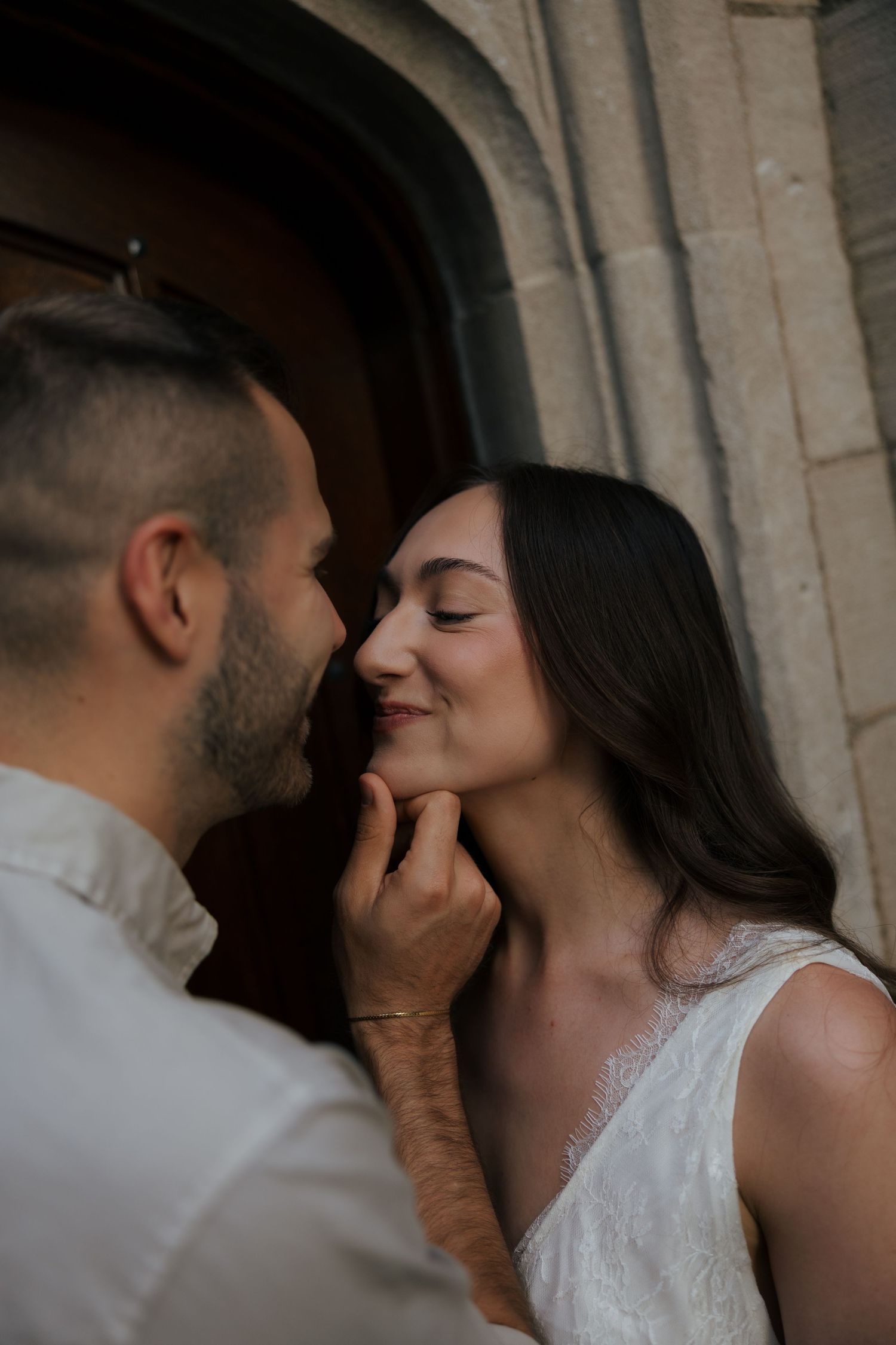 A groom tenderly holds his bride's chin as she smiles with closed eyes near a stone archway.