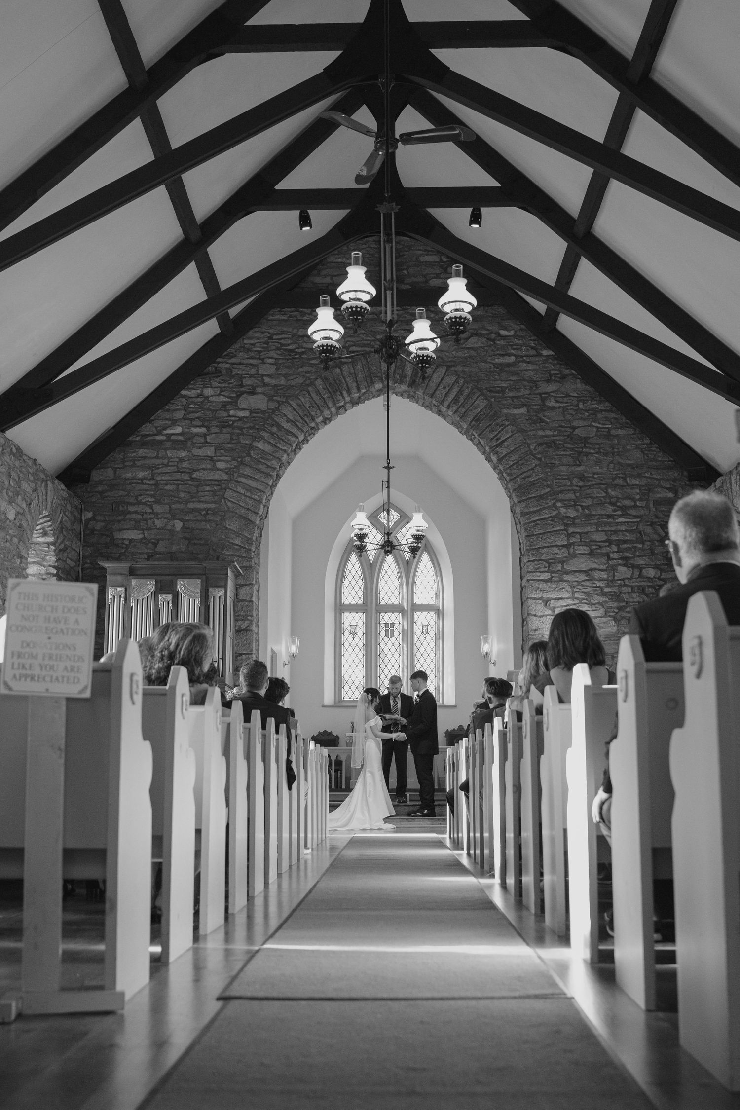 Black and white view down church aisle with exposed wooden beams and gothic arched windows at altar.
