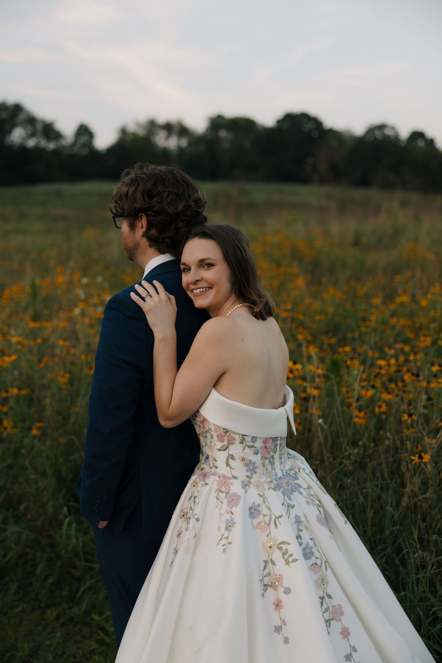 Smiling bride in floral ballgown embraces groom from behind in a sunny wildflower meadow at dusk.
