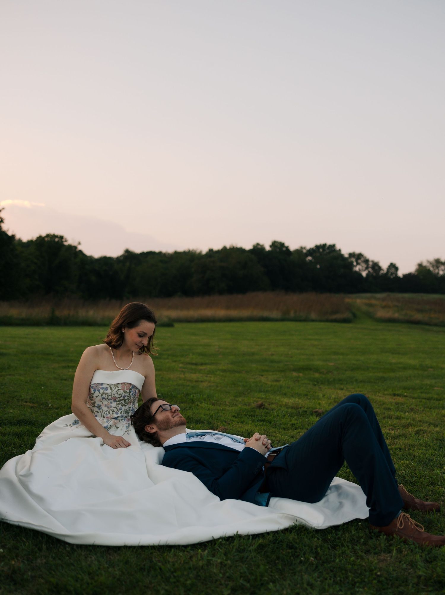 Bride and groom relax on grass at sunset, groom lying in bride's lap, sharing a joyful moment.
