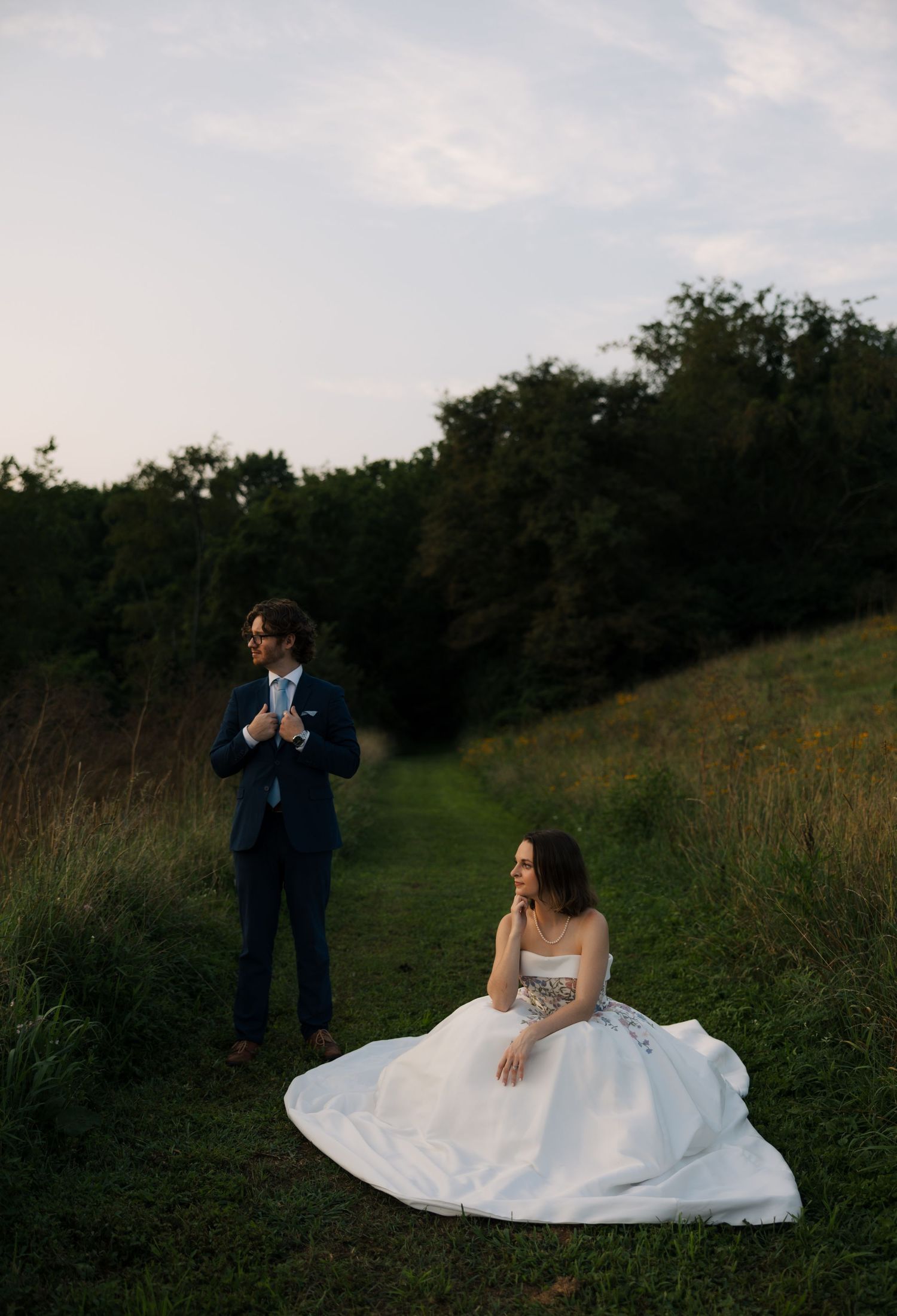Bride in white gown sits on grass path while groom in navy suit stands nearby at golden hour outdoor wedding.