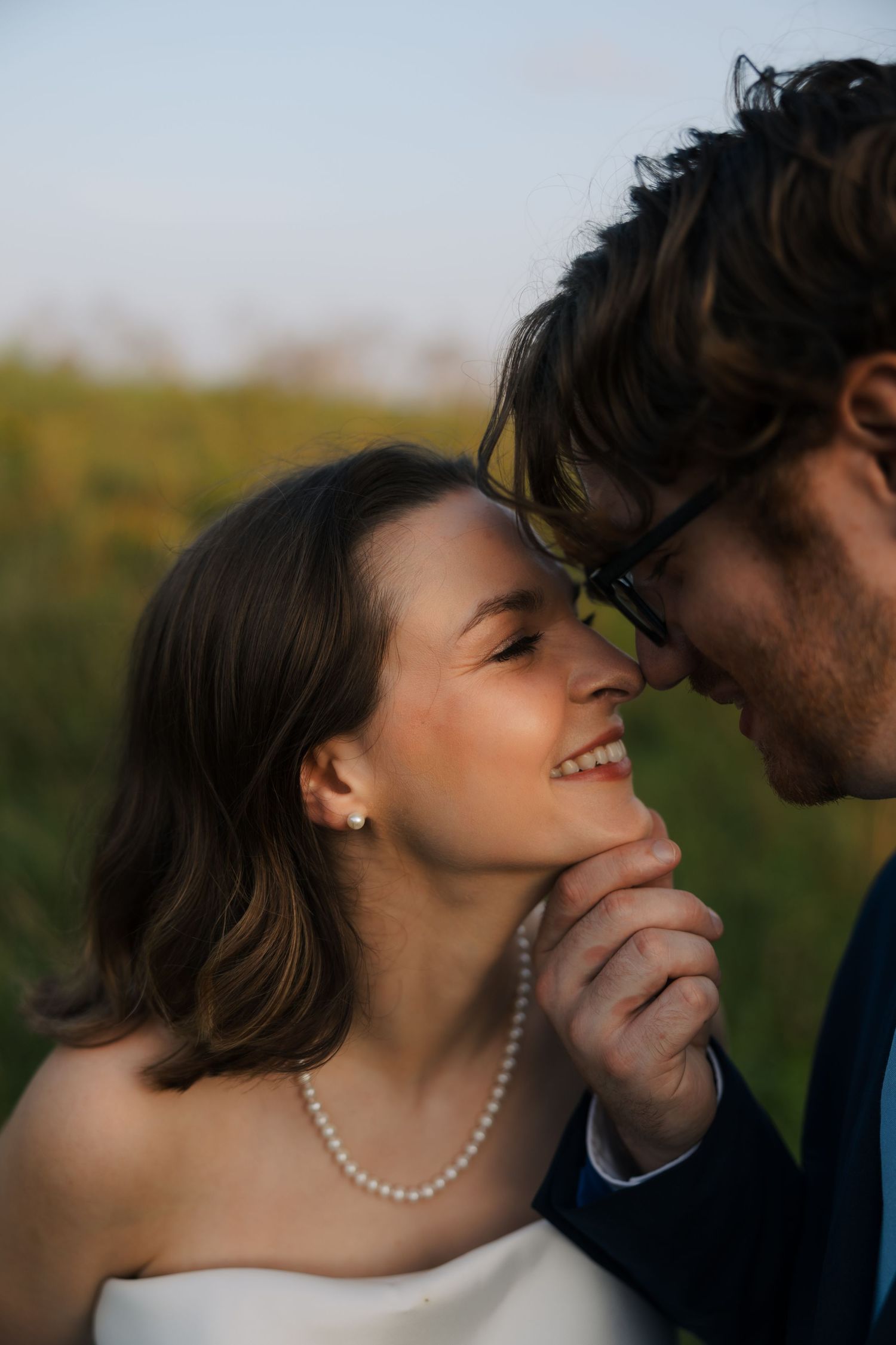 Smiling bride in pearl necklace nearly kisses groom wearing glasses in soft outdoor light.
