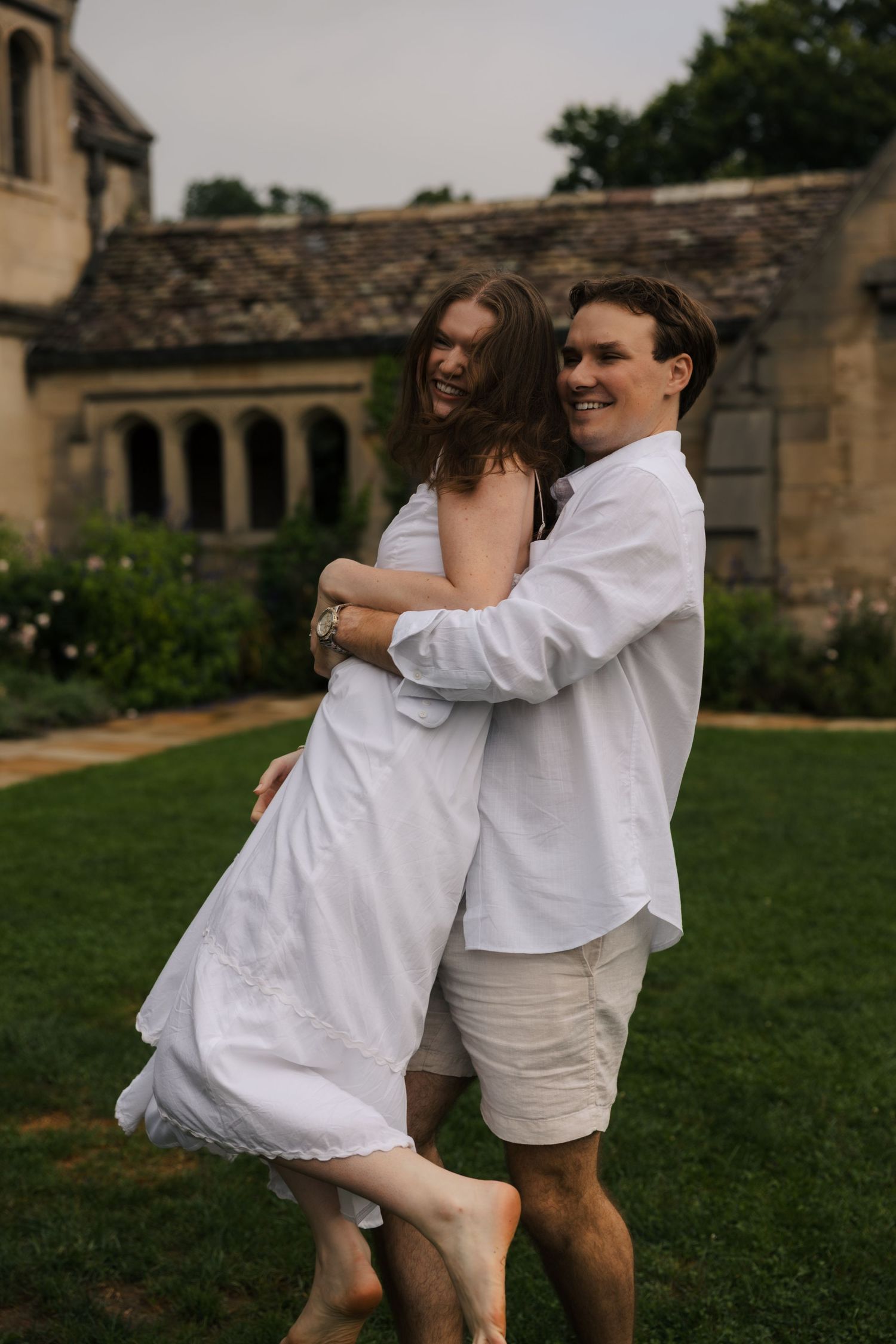 Couple in white outfits embrace and laugh together on green lawn beside historic stone manor.
