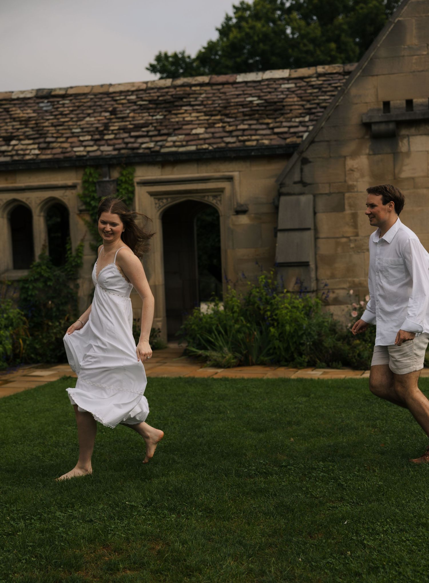 Laughing couple running barefoot across green grass in front of a charming stone cottage garden.