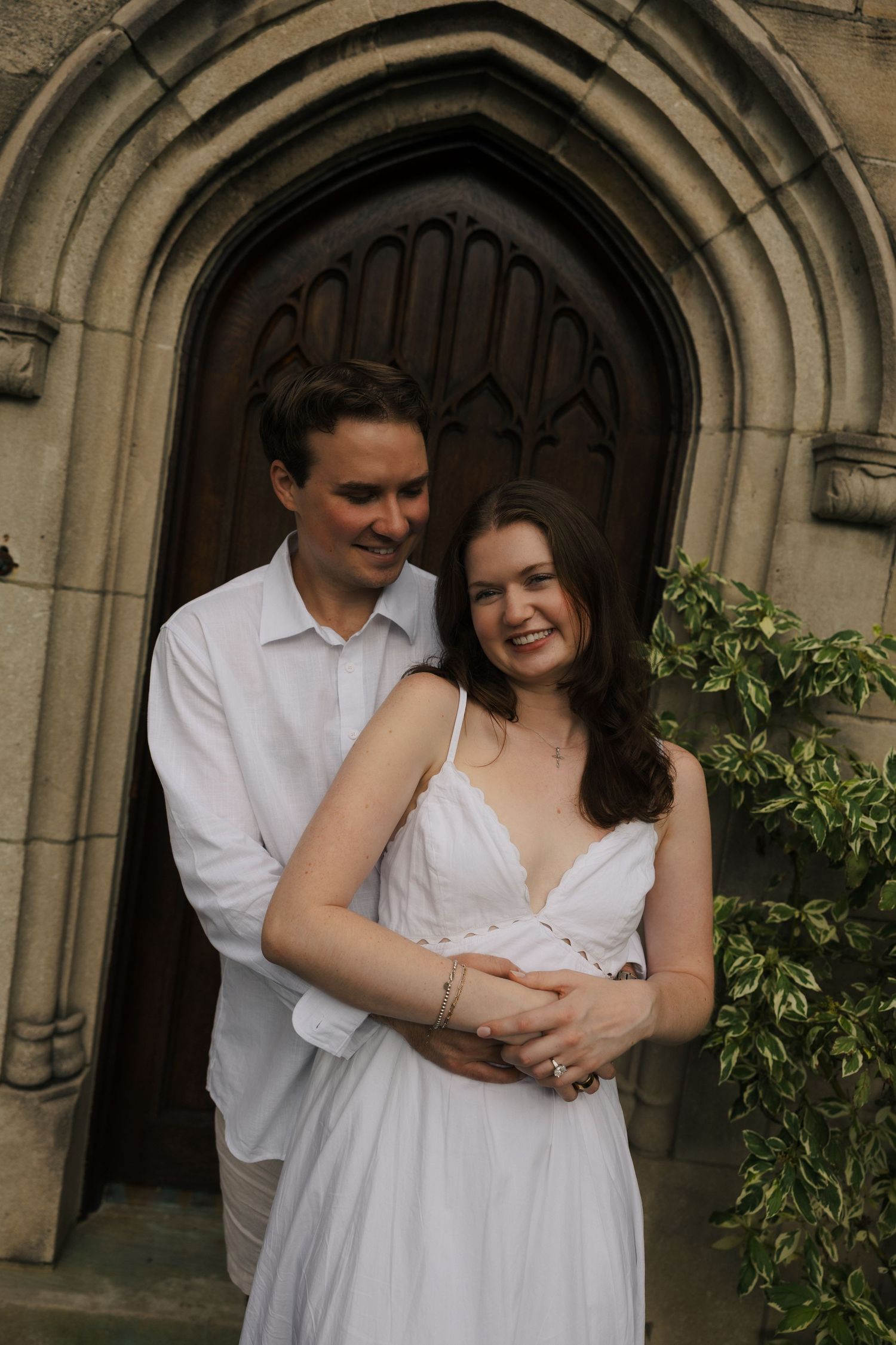 Smiling couple in white posing together before a grand arched wooden church door with lush greenery.