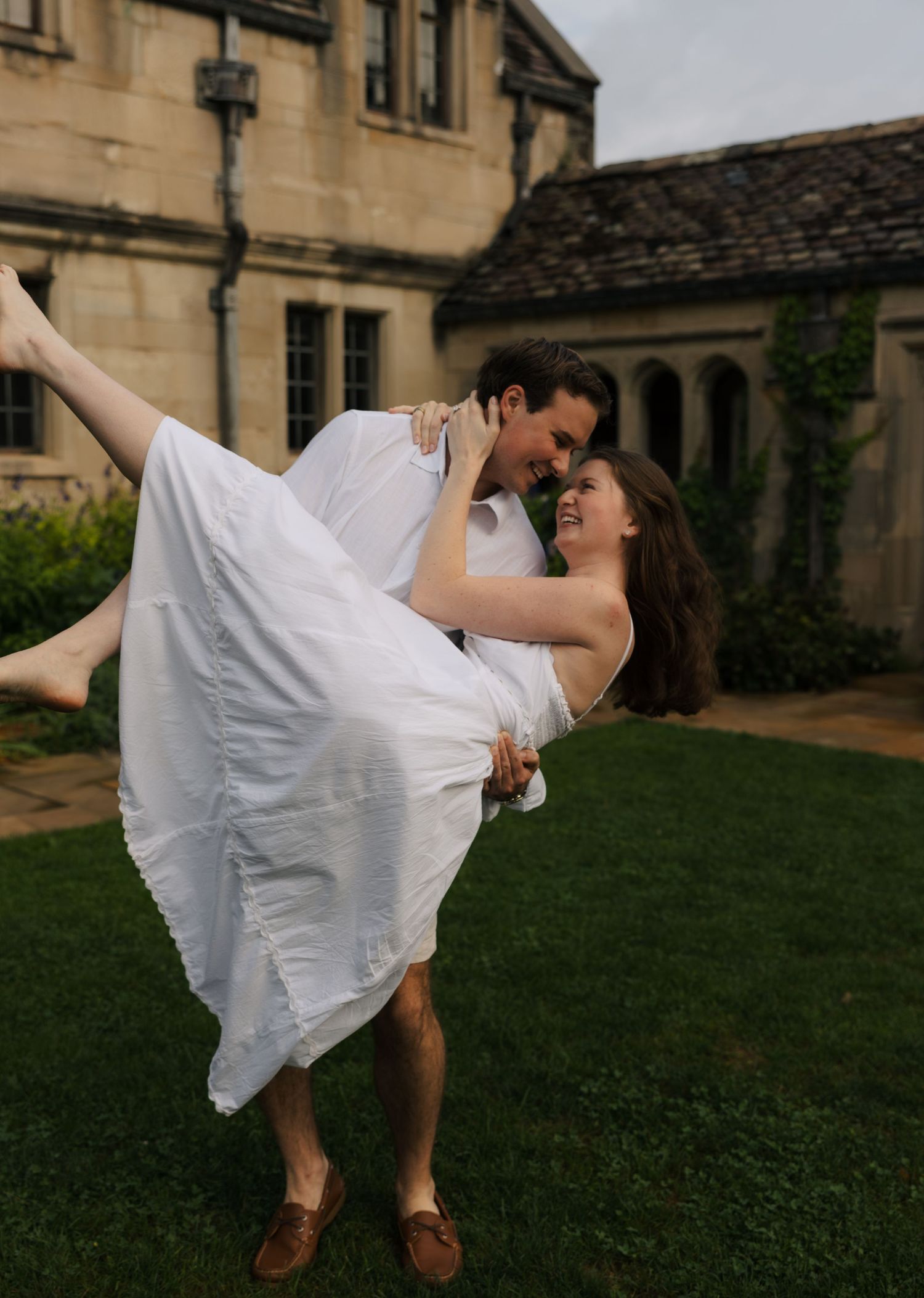 Man lifting woman in white dress outdoors, both laughing happily in front of a stone estate.