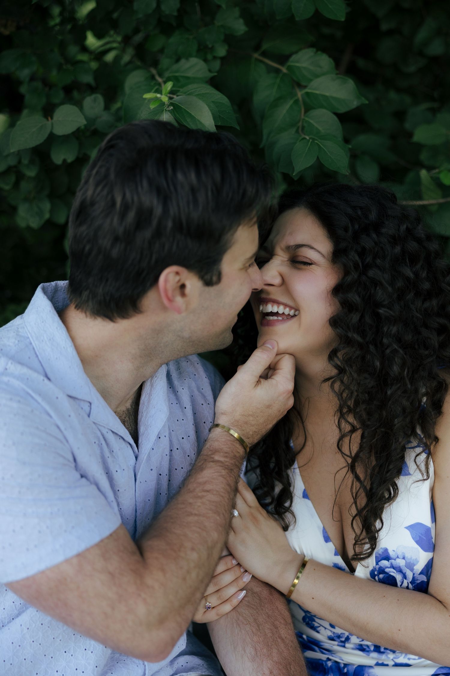 A couple shares a joyful, intimate moment laughing together in front of lush green foliage.