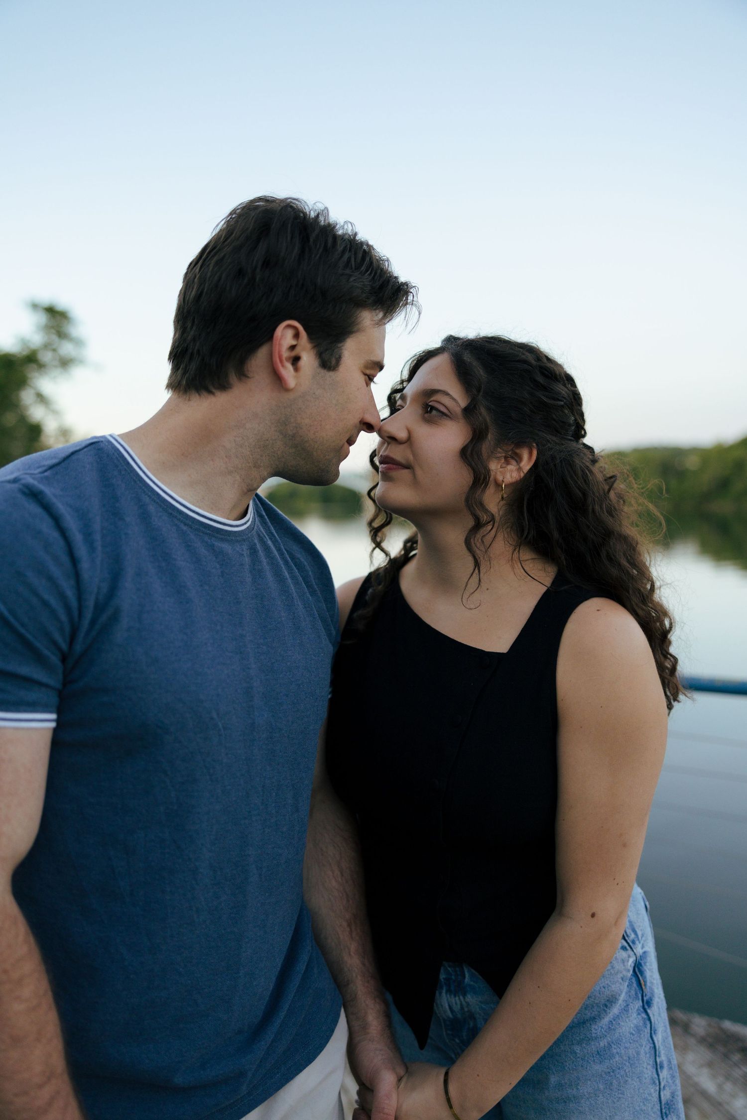 Couple sharing a romantic moment by a river at dusk, man in blue tee and woman in black top holding hands.