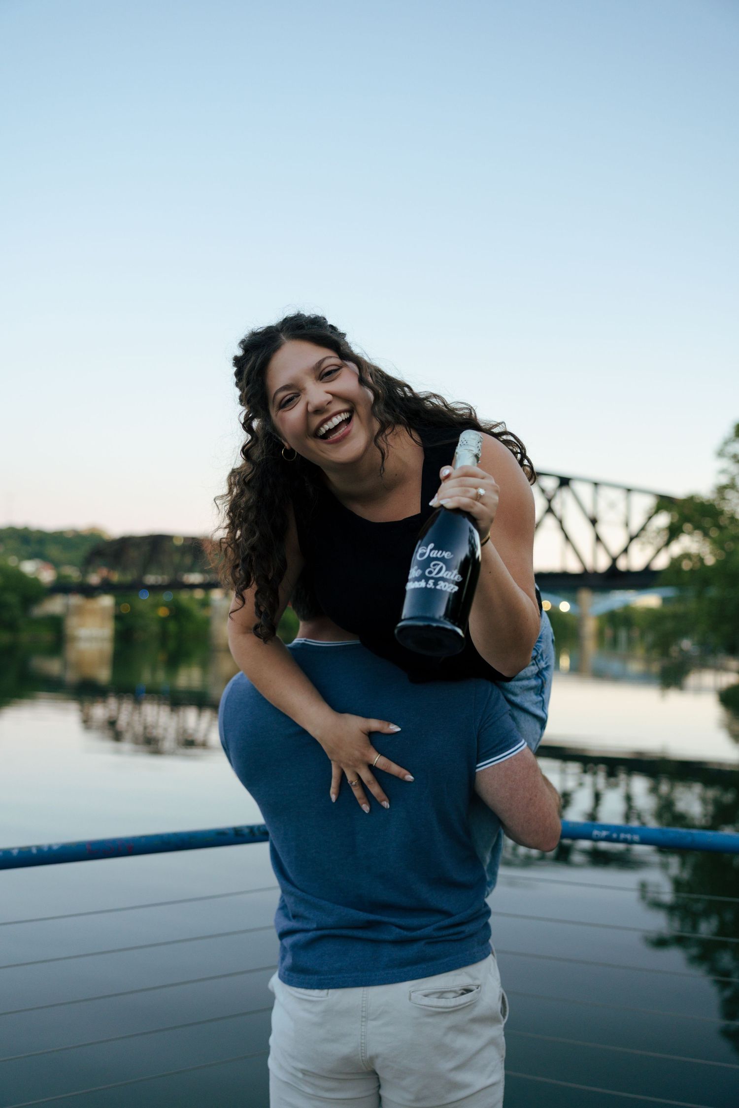 A joyful woman holds a champagne bottle while being lifted by her partner on a waterfront bridge at dusk.