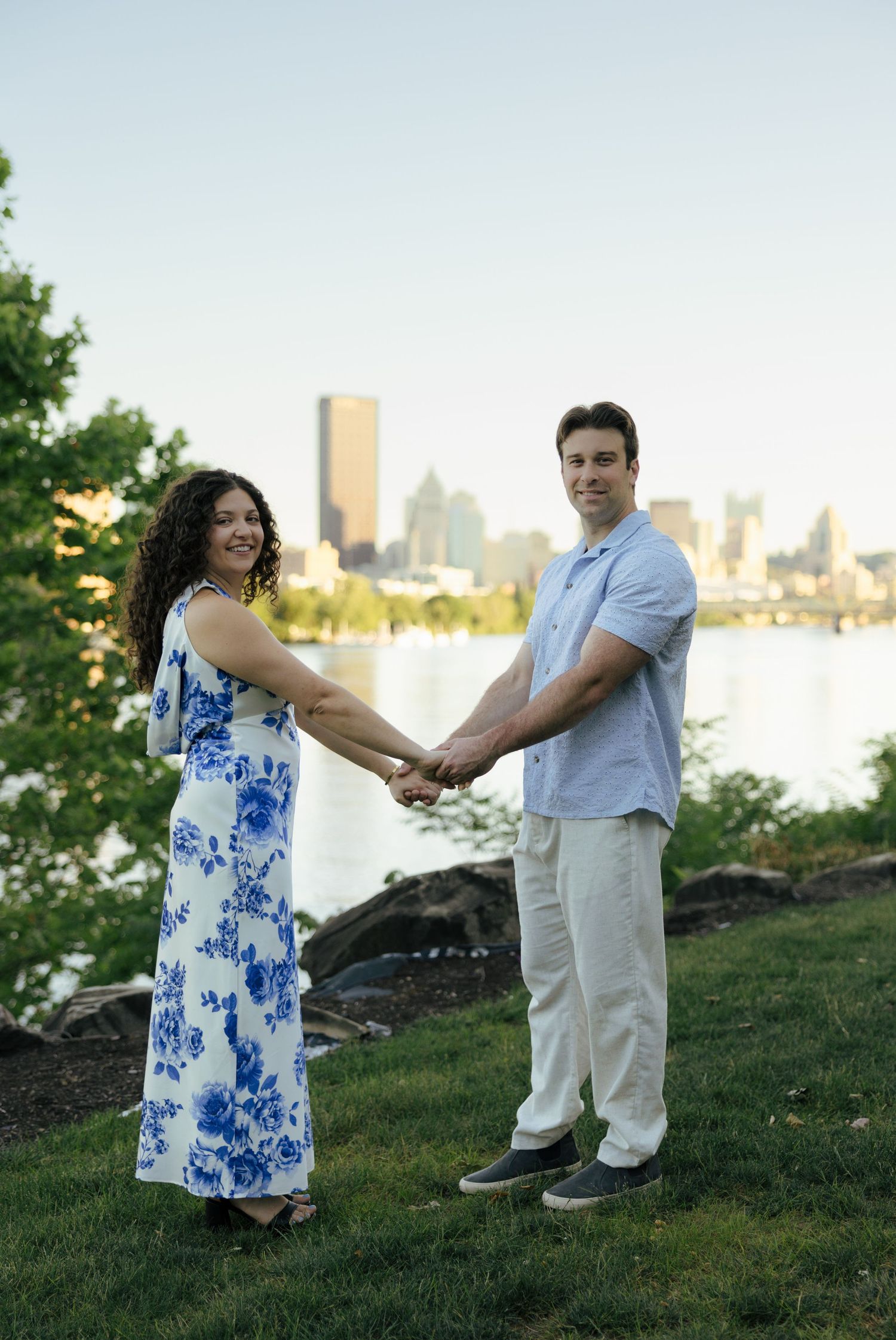 Smiling couple holding hands facing camera with city skyline and river in the background at dusk.