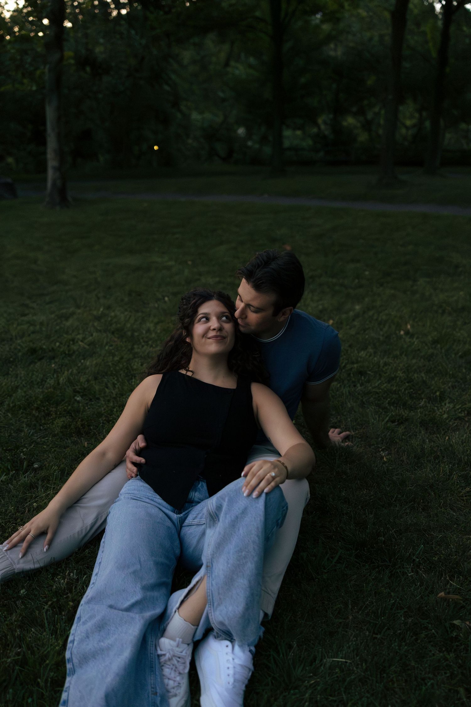 A couple sits together on grass in a park at dusk, sharing a tender romantic moment.