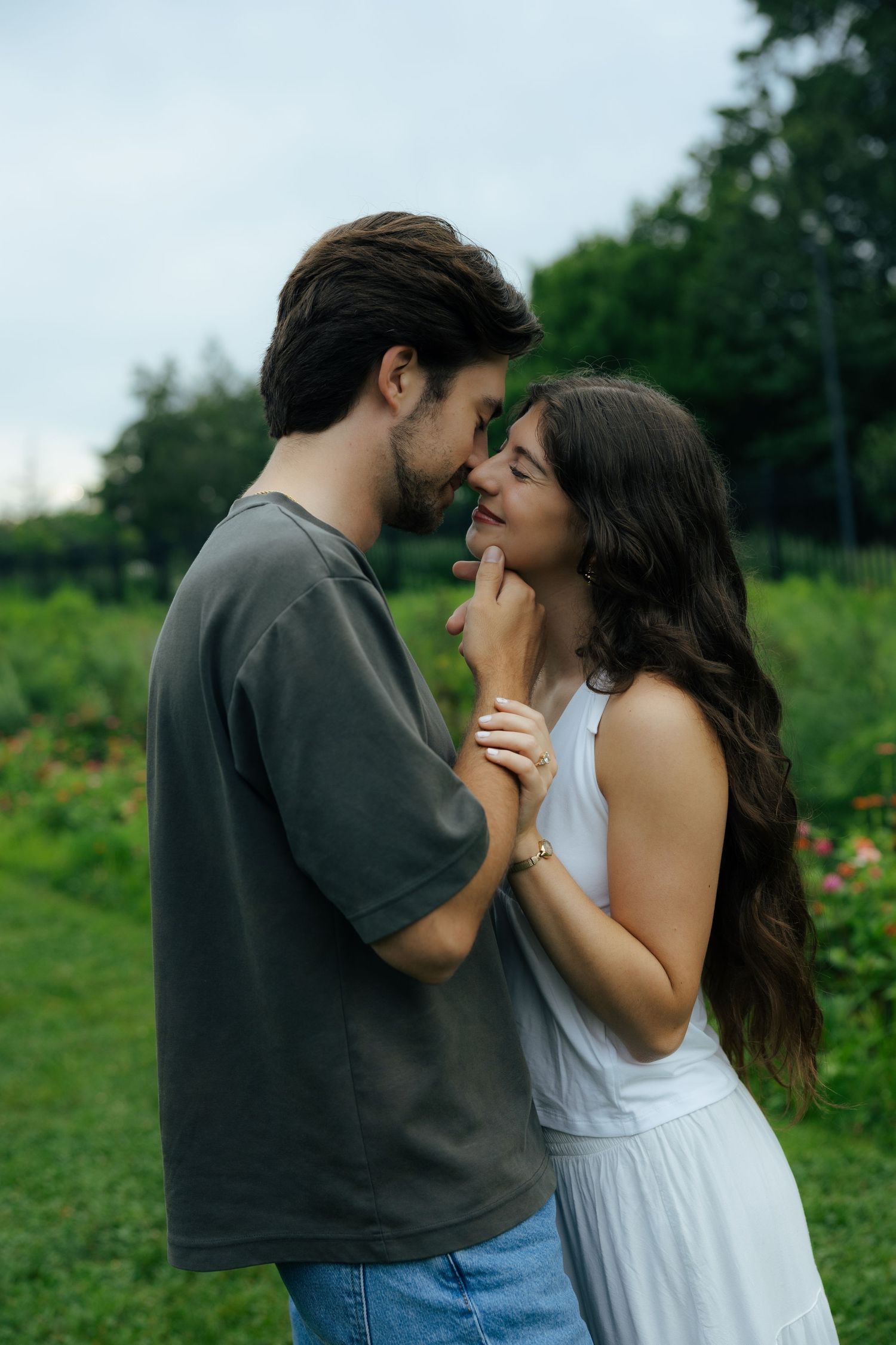 A romantic couple nearly kissing in a lush green garden, woman in white dress, man in gray t-shirt.