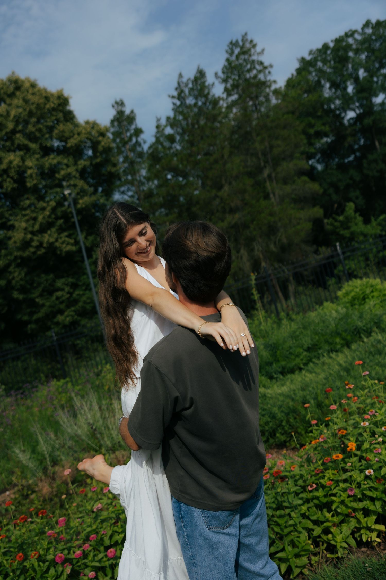 A couple embraces and smiles in a lush garden, the woman in a white dress wrapping her arms around a man in a gray shirt.