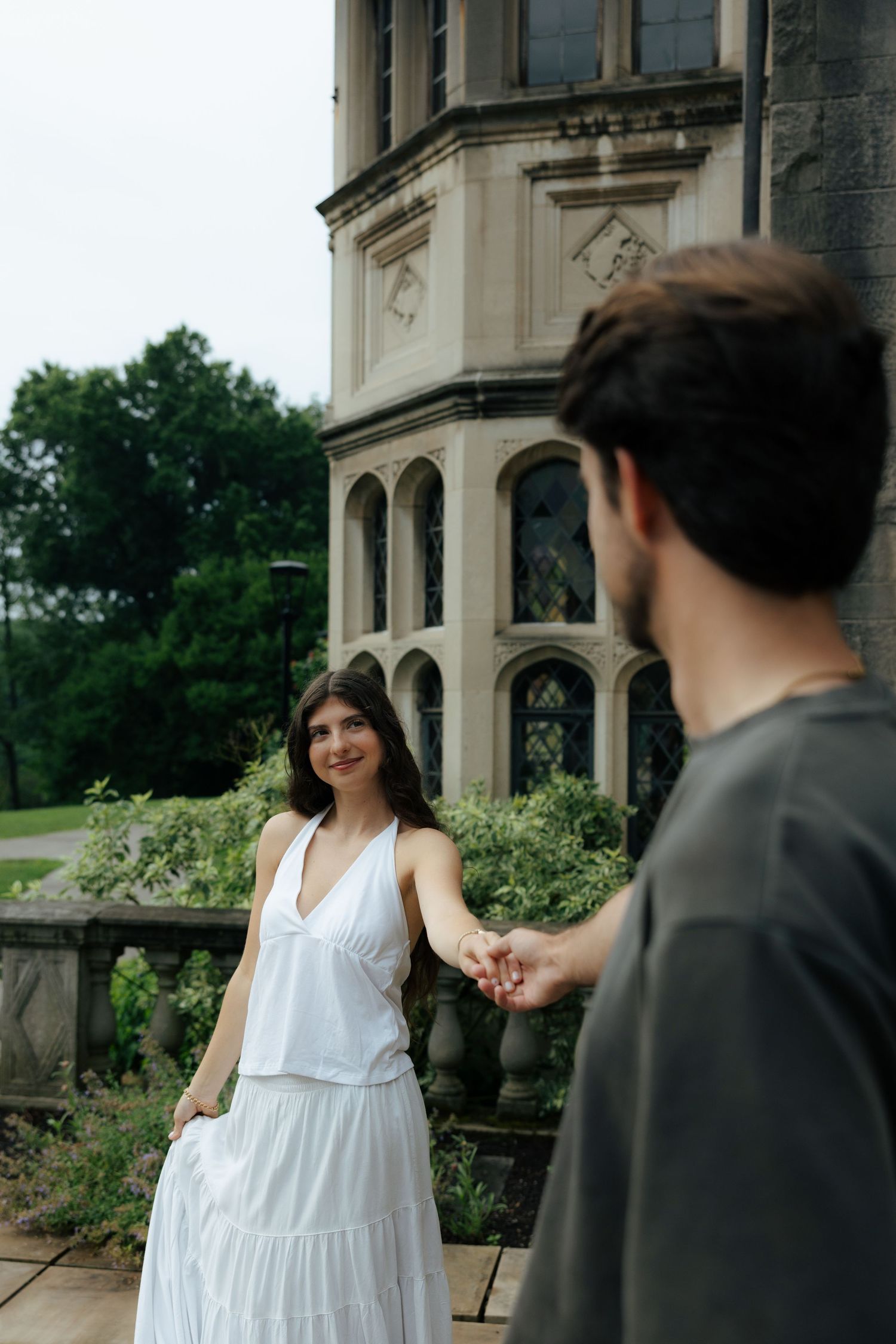 A smiling woman in a white dress holds hands with a man near a historic stone building surrounded by greenery.