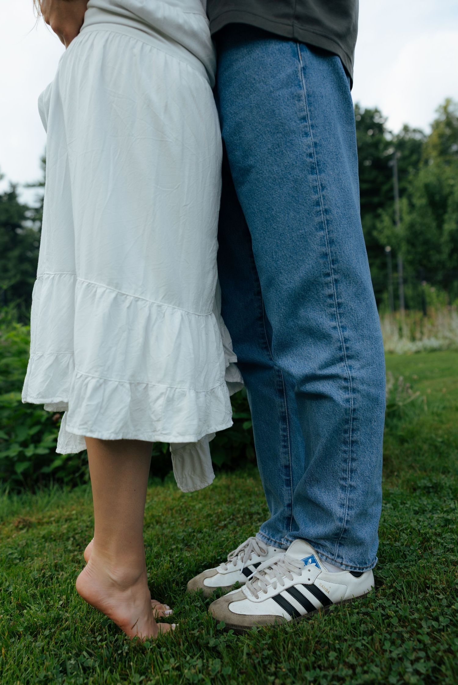 Close-up of a couple standing together, the woman barefoot in a white dress beside a man in jeans and Adidas sneakers.