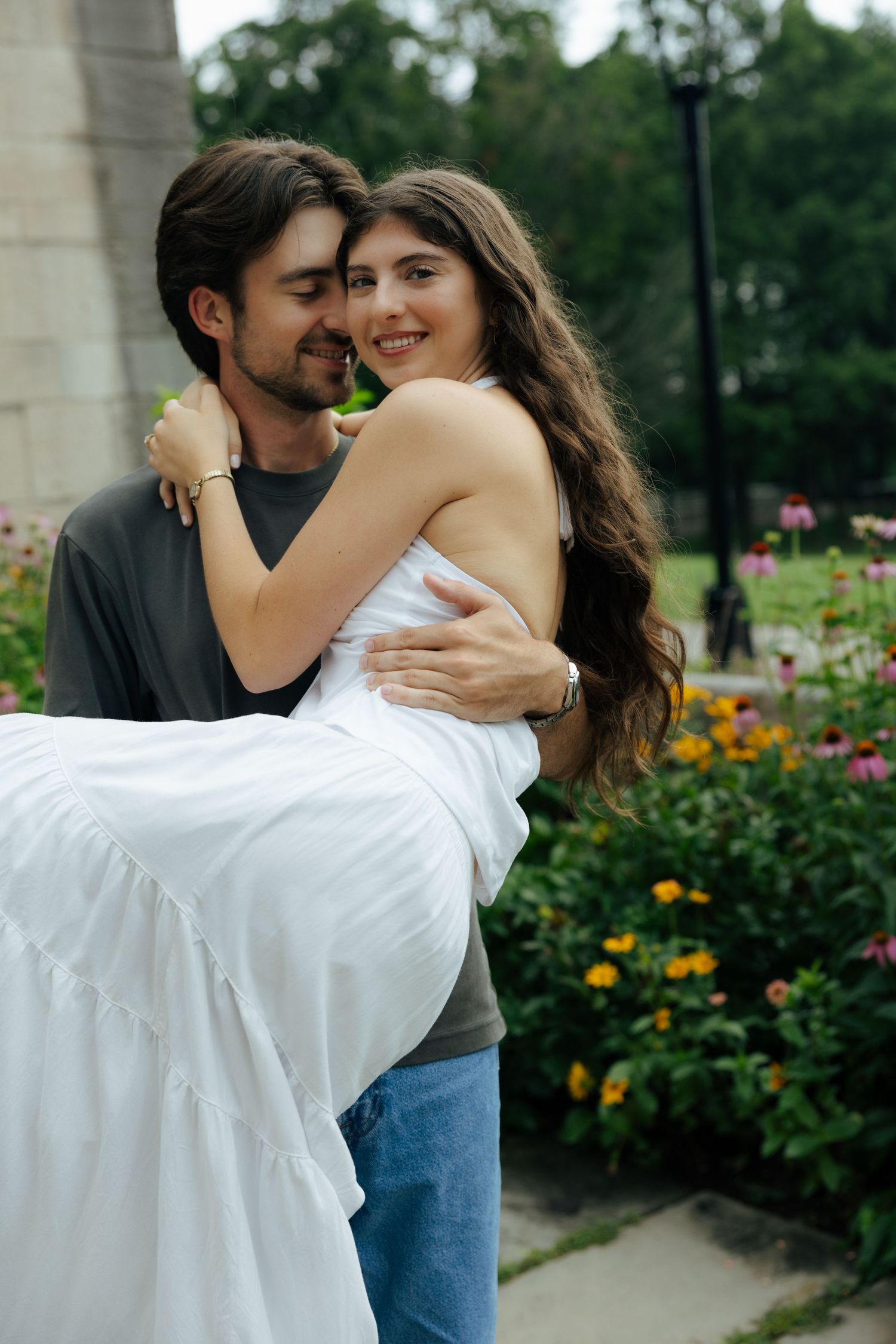 A man carries a smiling woman in a white dress in a colorful garden.