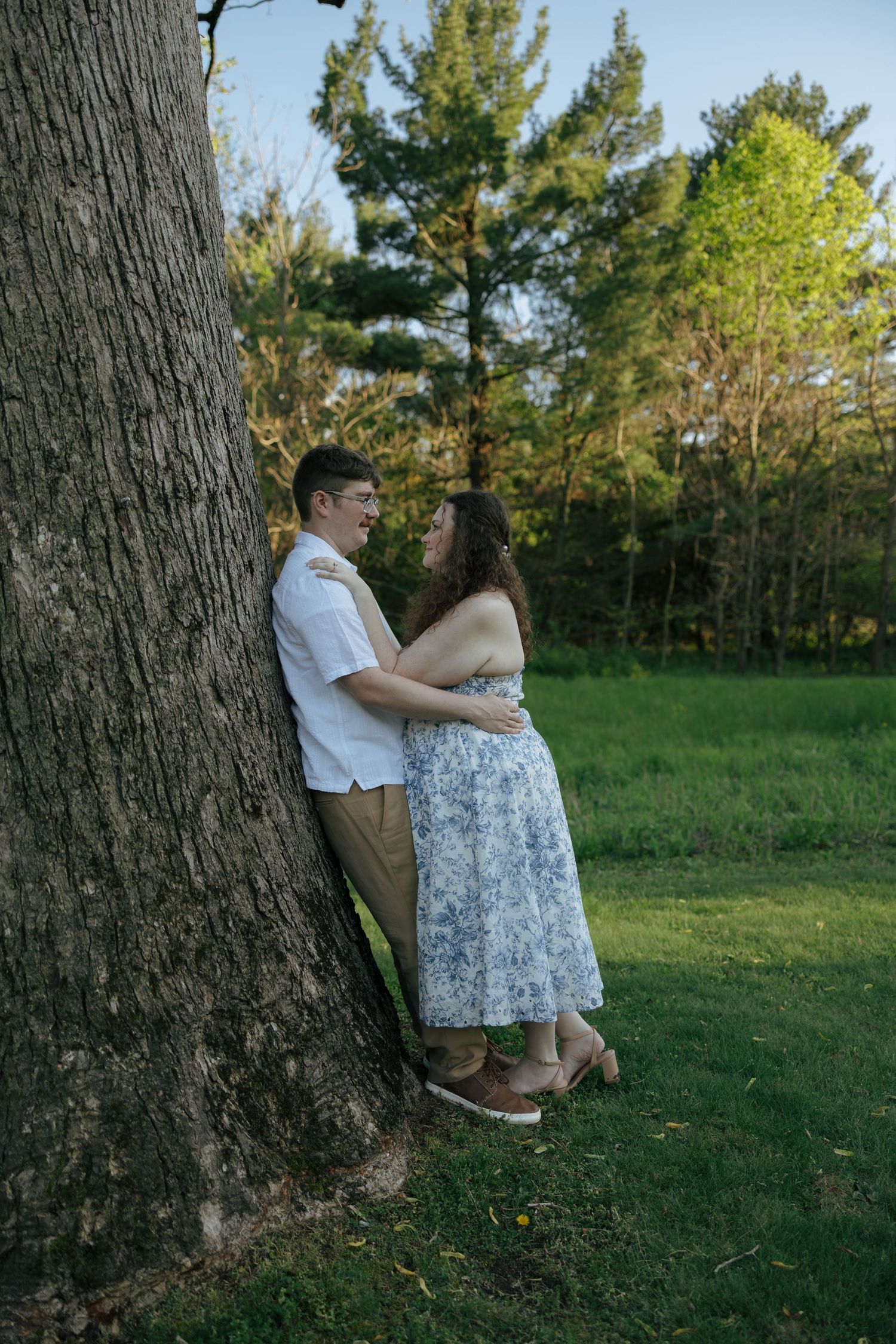 Couple sharing a tender moment leaning against a large tree trunk in a green meadow at dusk.