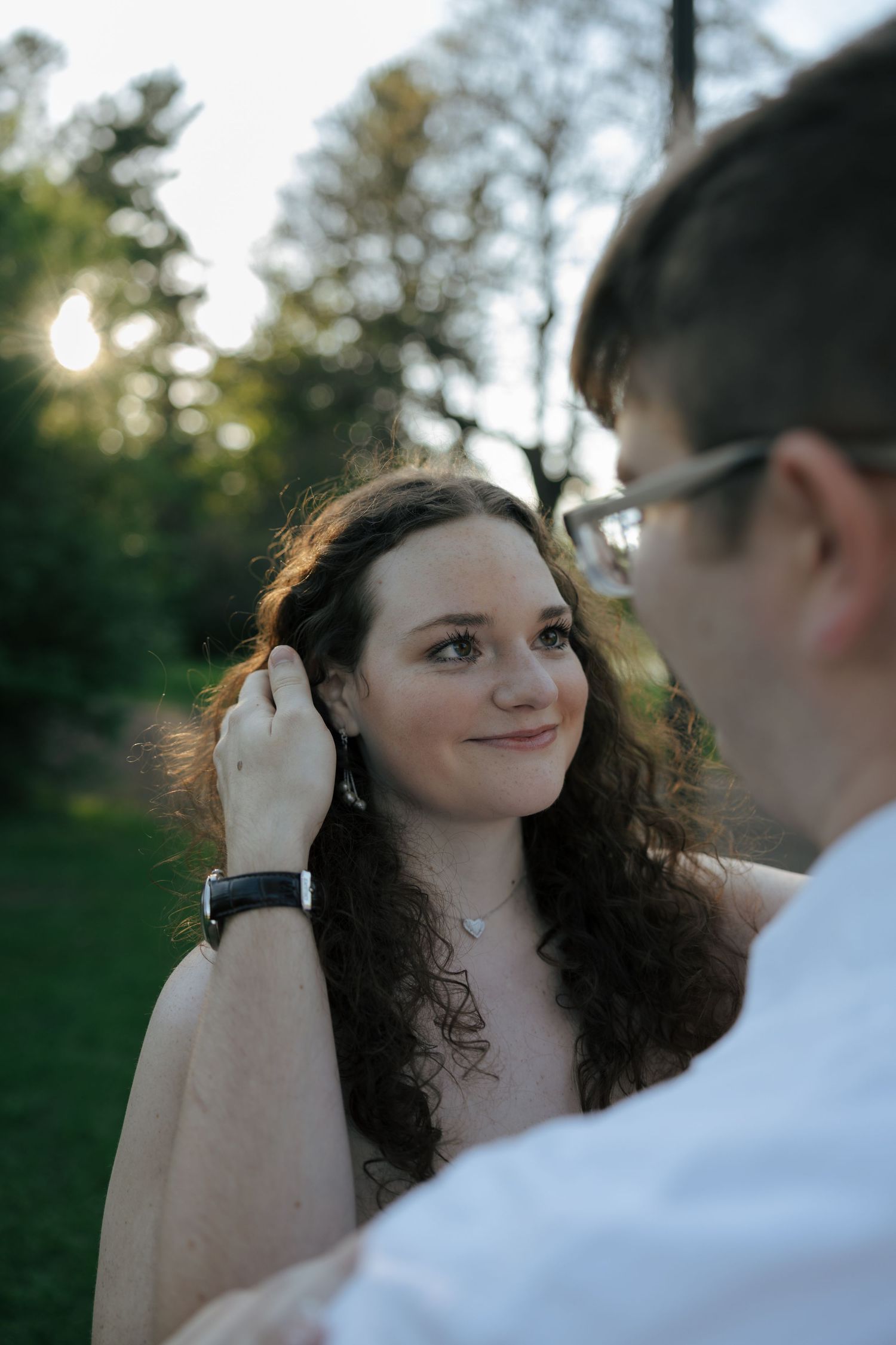 Man gently tucking hair behind woman's ear as she smiles warmly in a sunlit park.
